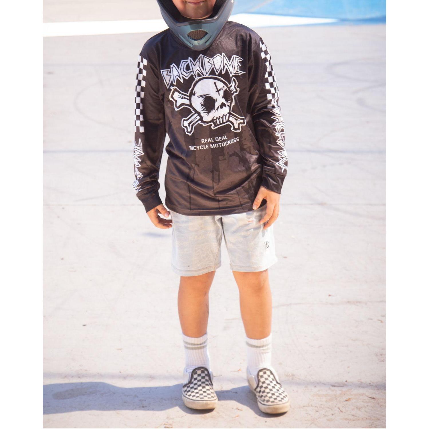 A child in a helmet, Backbone 2026 Gnar Drip Jersey with skull graphic, light shorts, white socks, and checkered slip-on shoes stands on a concrete surface.