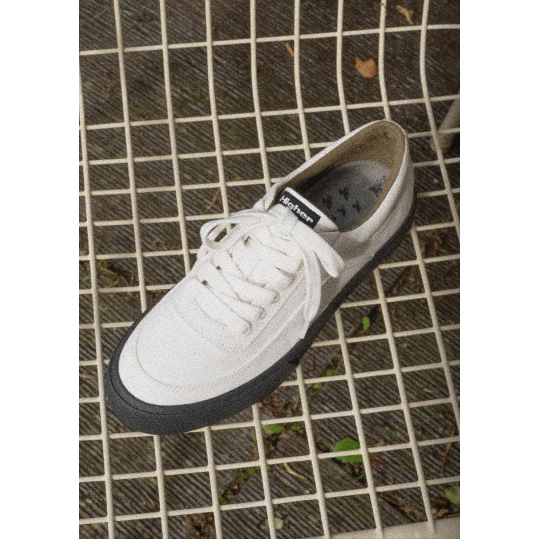 The Higher Crypt Canvas Lace Up Shoes (Raw Ecru/Black) feature a white upper, black grippy sole, and white laces, displayed on a white metal grid chair from an overhead view.