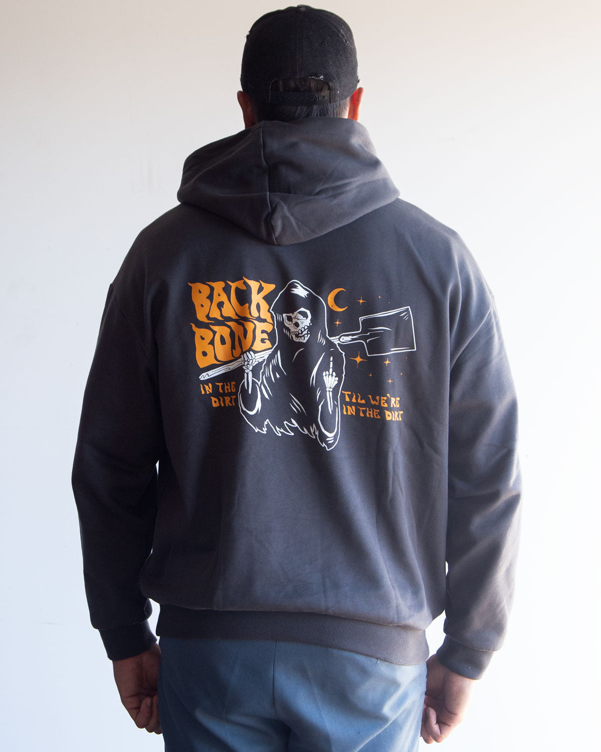 A person wearing the Backbone In the Dirt Hoodie—a heavyweight, oversized hoodie featuring a graphic of a skeleton and moon, with the text “BACK BONE IN THE DIRT ALL WEEK IN THE DIRT” printed on the back.