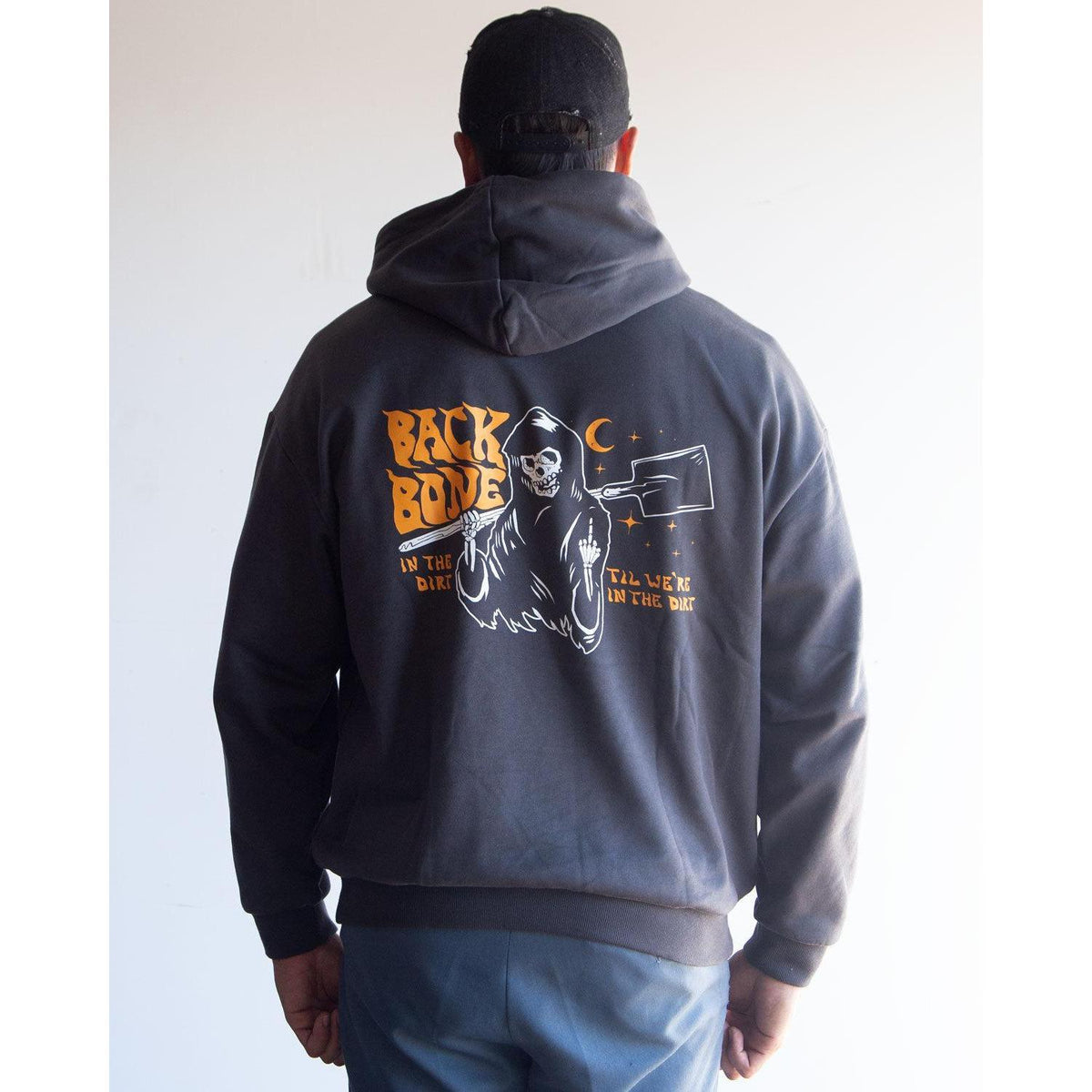A person wearing the Backbone In the Dirt Hoodie—a heavyweight, oversized hoodie featuring a graphic of a skeleton and moon, with the text “BACK BONE IN THE DIRT ALL WEEK IN THE DIRT” printed on the back.