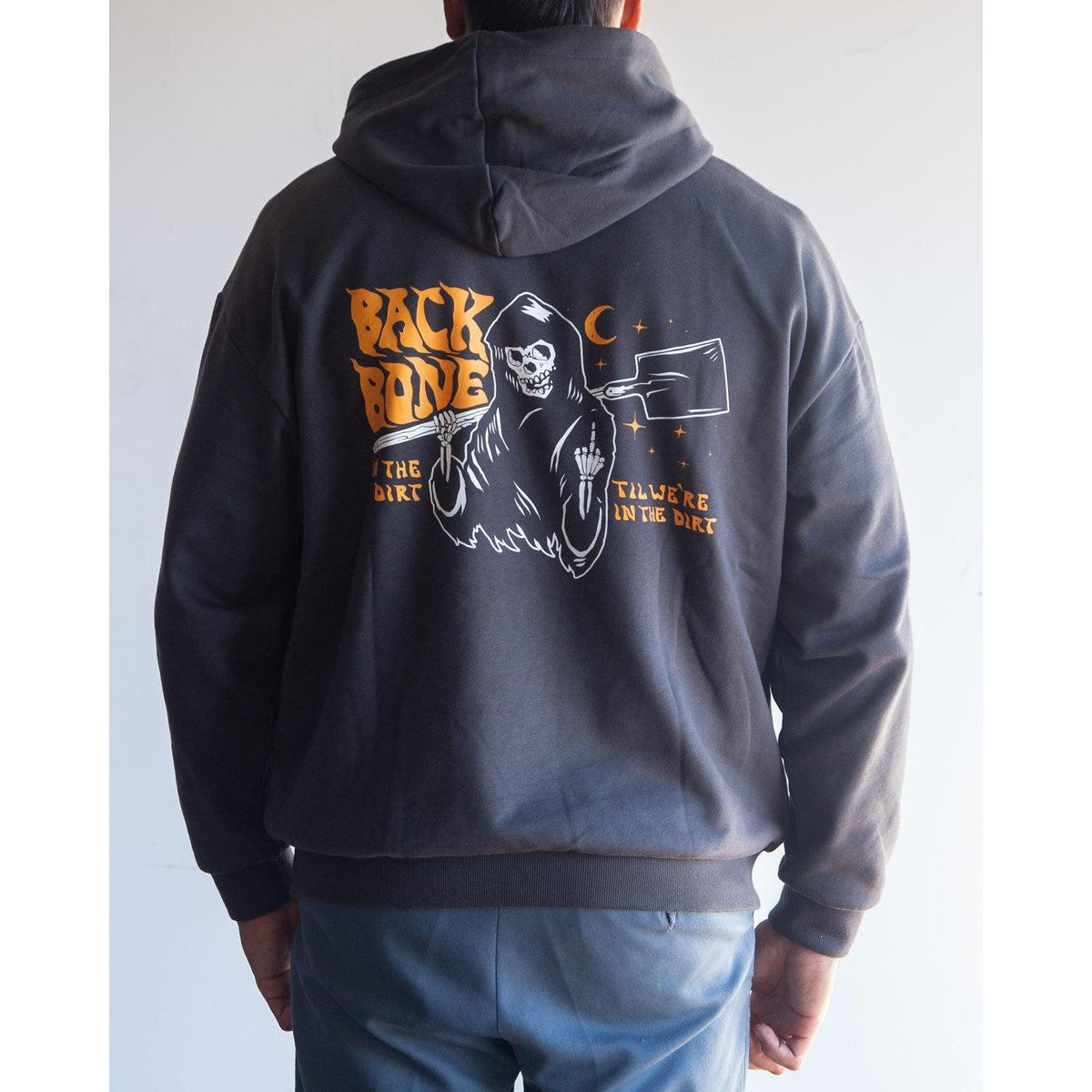 A person is wearing the Backbone In the Dirt Hoodie, an oversized black hoodie featuring a Grim Reaper graphic holding a cleaver, with “BACKBONE” and “I’ll Work In The Dirt” text on the back.