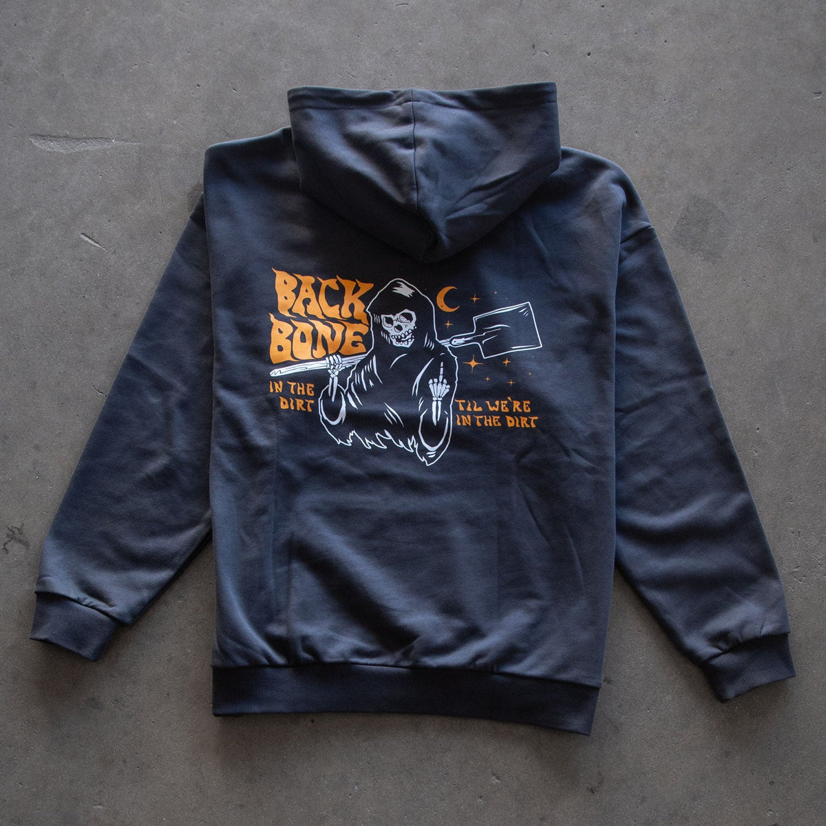 A Backbone In the Dirt Hoodie in black heavy weight fabric is shown lying flat, featuring a graphic of a grim reaper holding a scythe with orange text that says “BACK BONE” and “TIL WE’RE IN THE DIRT.”.
