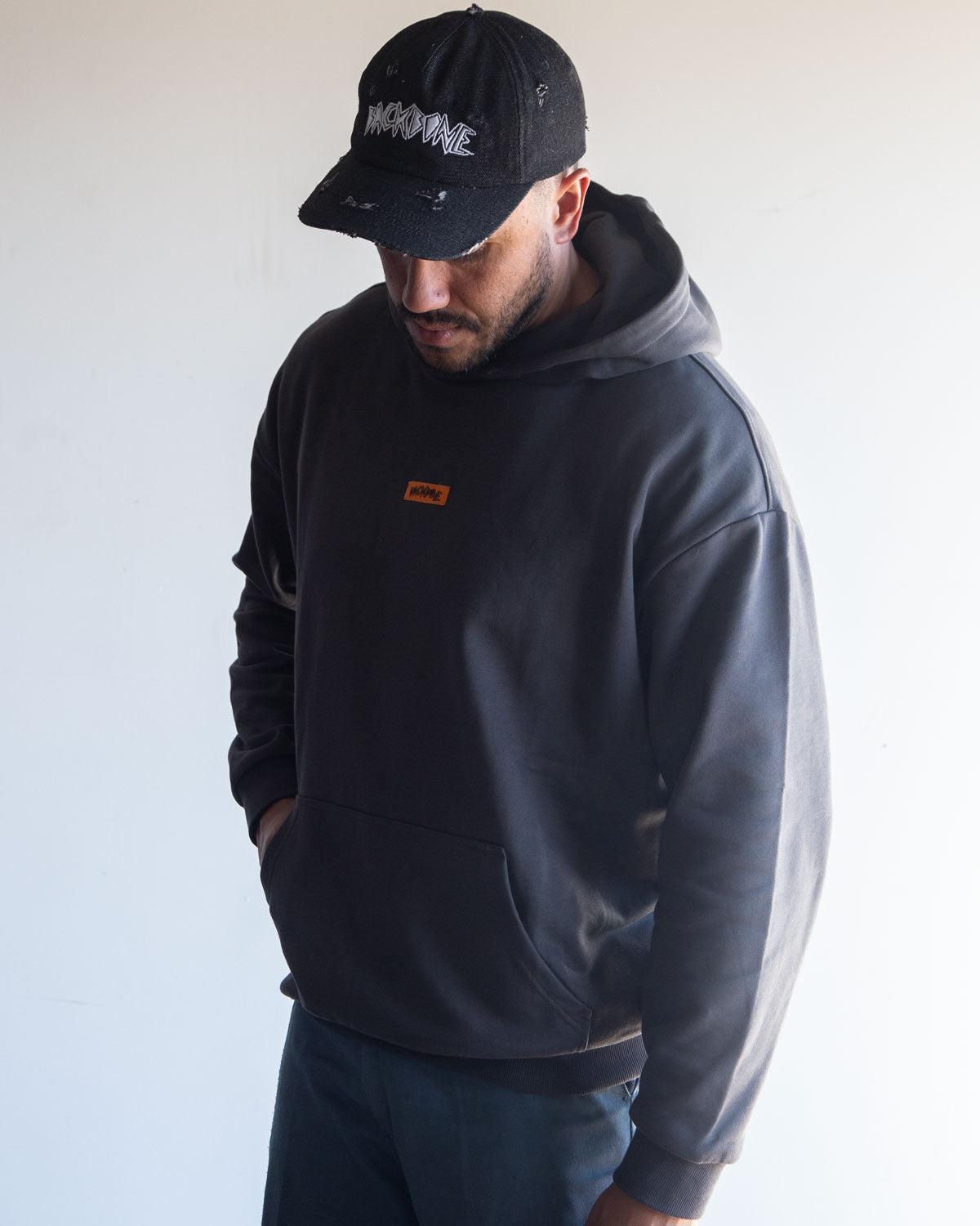 A man wearing the Backbone In the Dirt Hoodie—a black, heavyweight style with a small orange label—paired with a black cap featuring white text, stands against a plain light background looking down.