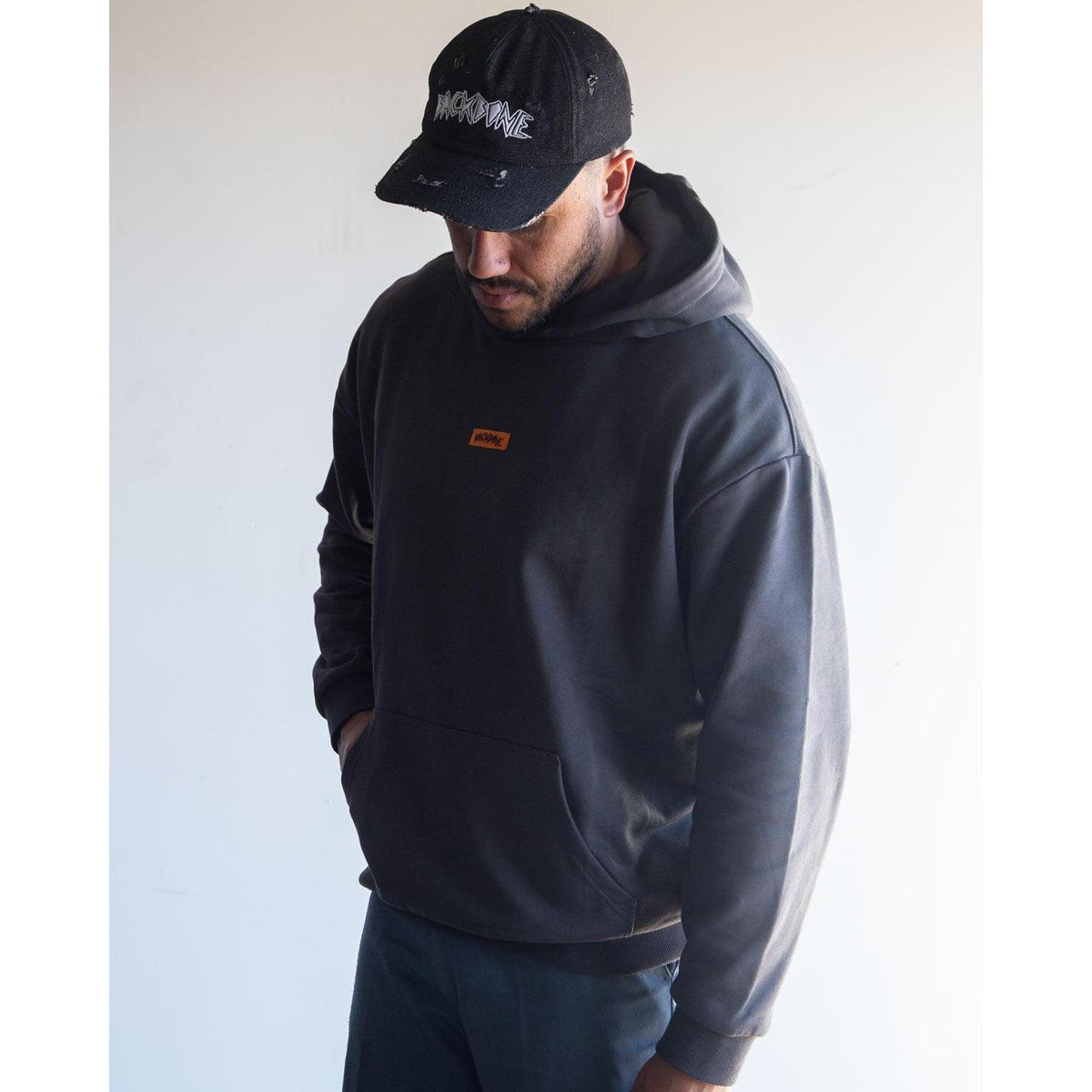 A man wearing the Backbone In the Dirt Hoodie—a black, heavyweight style with a small orange label—paired with a black cap featuring white text, stands against a plain light background looking down.