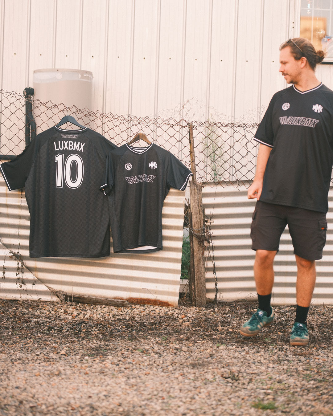 A person stands outdoors by a fence with two black sports jerseys hanging on it, including the LUXBMX MMX Soccer Jersey - Custom Back Print.