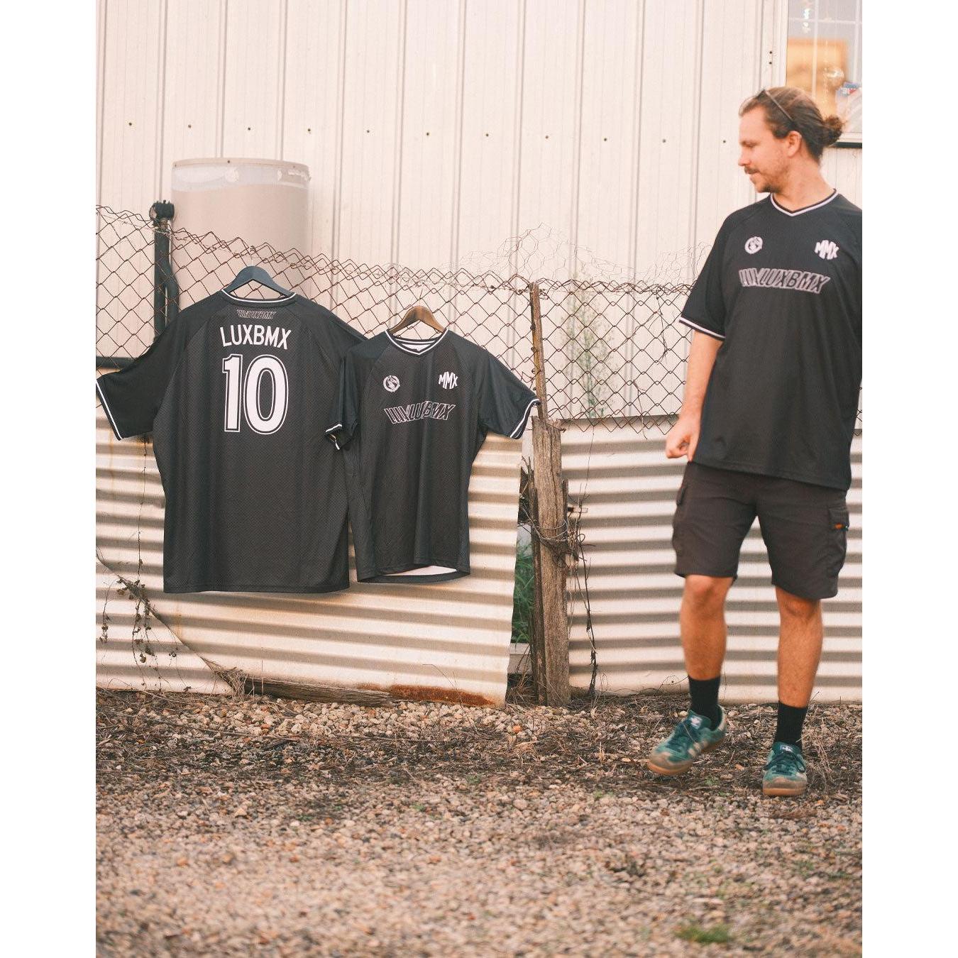 A person stands outdoors by a fence with two black sports jerseys hanging on it, including the LUXBMX MMX Soccer Jersey - Custom Back Print.