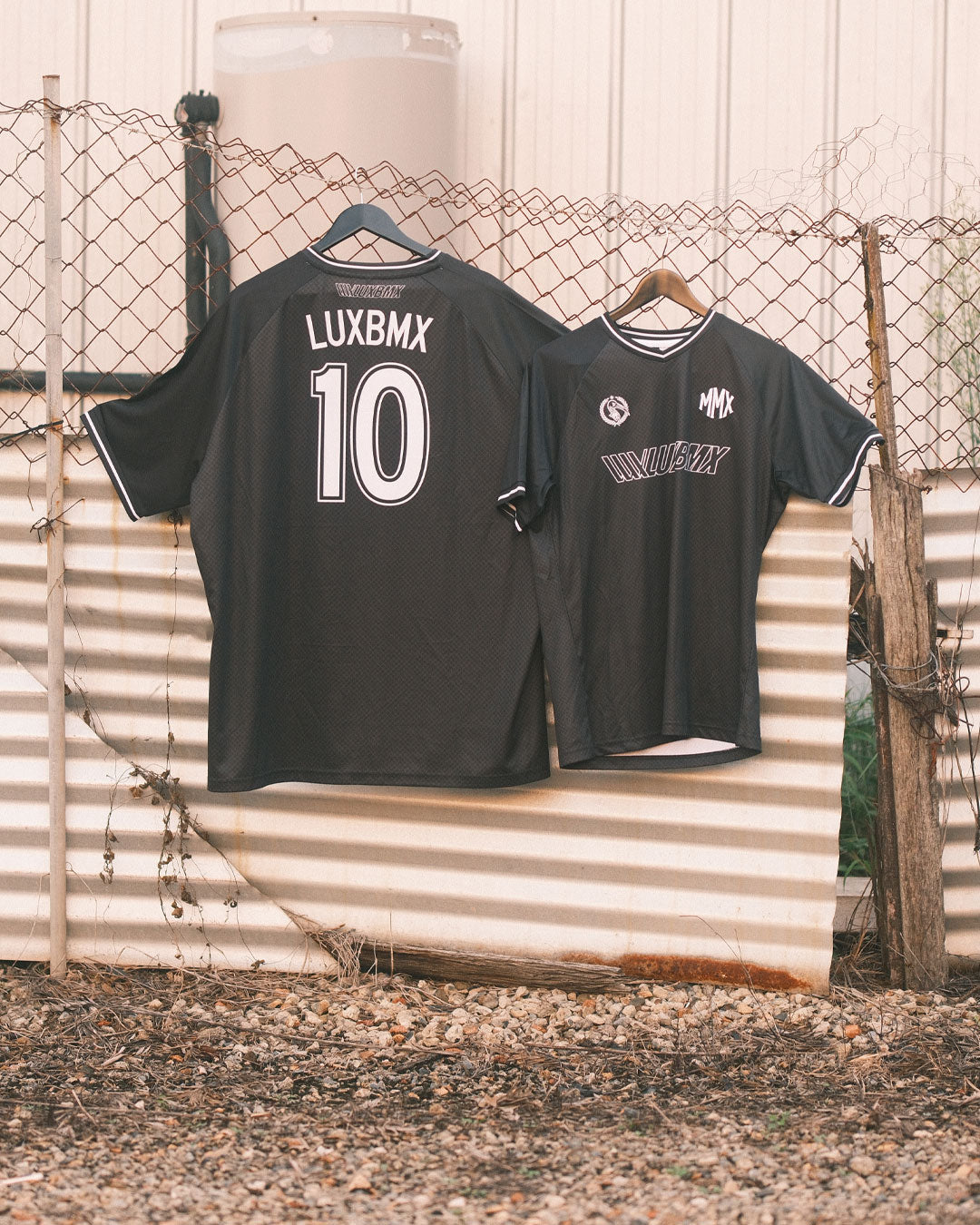 Two black LUXBMX MMX Soccer Jerseys with custom back print hang on a chain-link fence outdoors, highlighting the comfort and style of this breathable bike jersey.