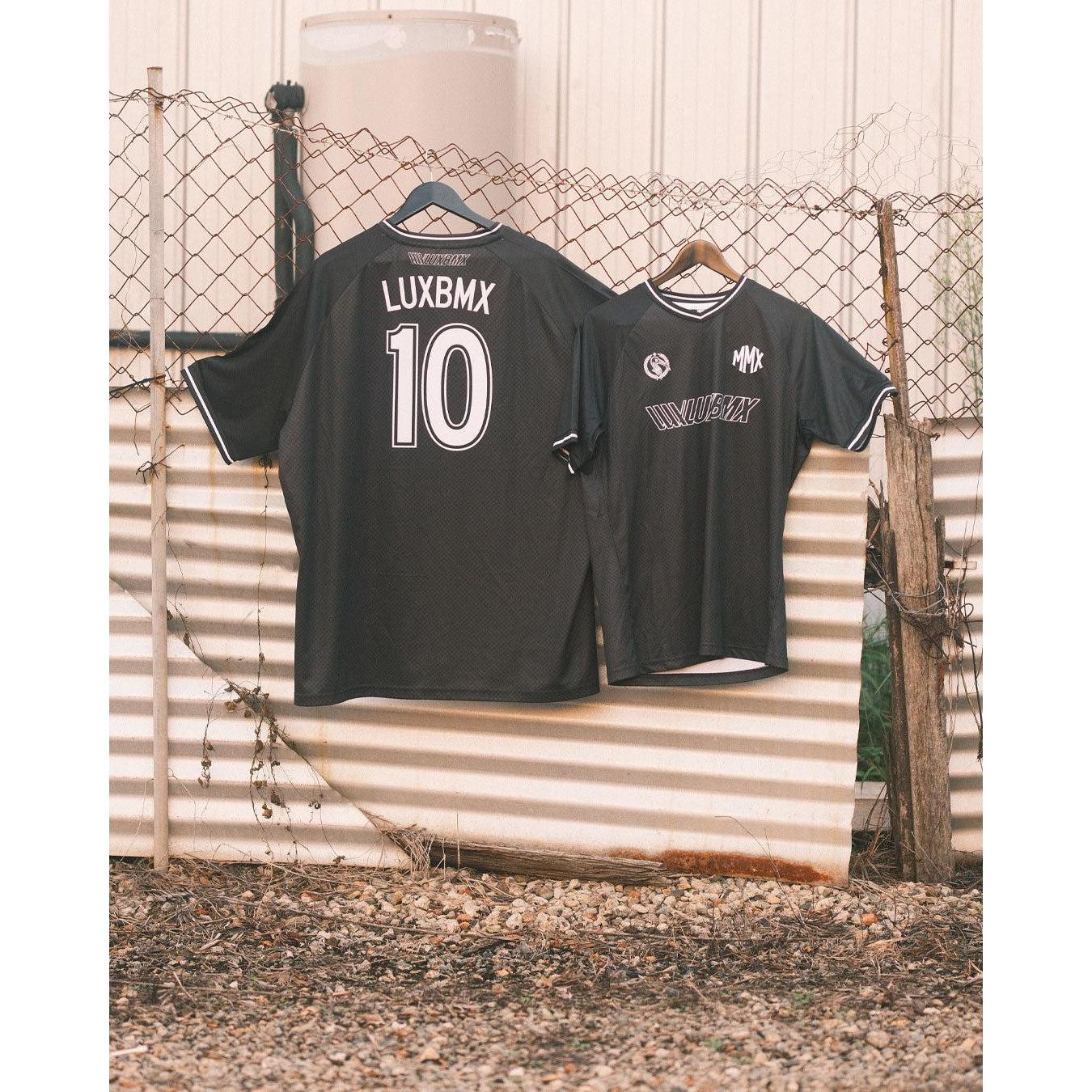 Two black LUXBMX MMX Soccer Jerseys with custom back print hang on a chain-link fence outdoors, highlighting the comfort and style of this breathable bike jersey.