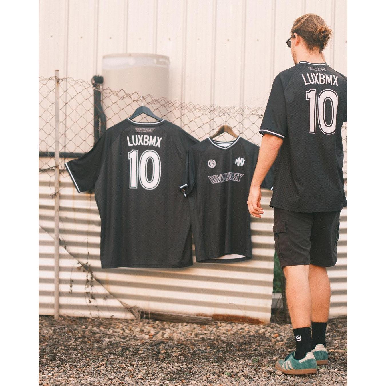A person in a black LUXBMX MMX Soccer Jersey - Custom Back Print stands outdoors near two identical jerseys hanging on a metal fence.