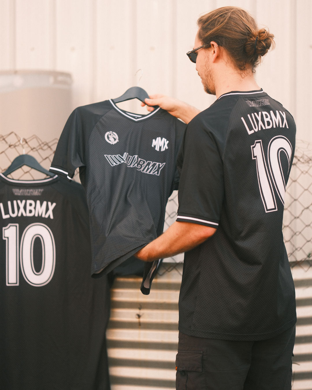 A man in sunglasses holds a black LUXBMX MMX Soccer Jersey with a custom back print (number 10), standing next to two similar breathable bike jerseys hanging on a fence.