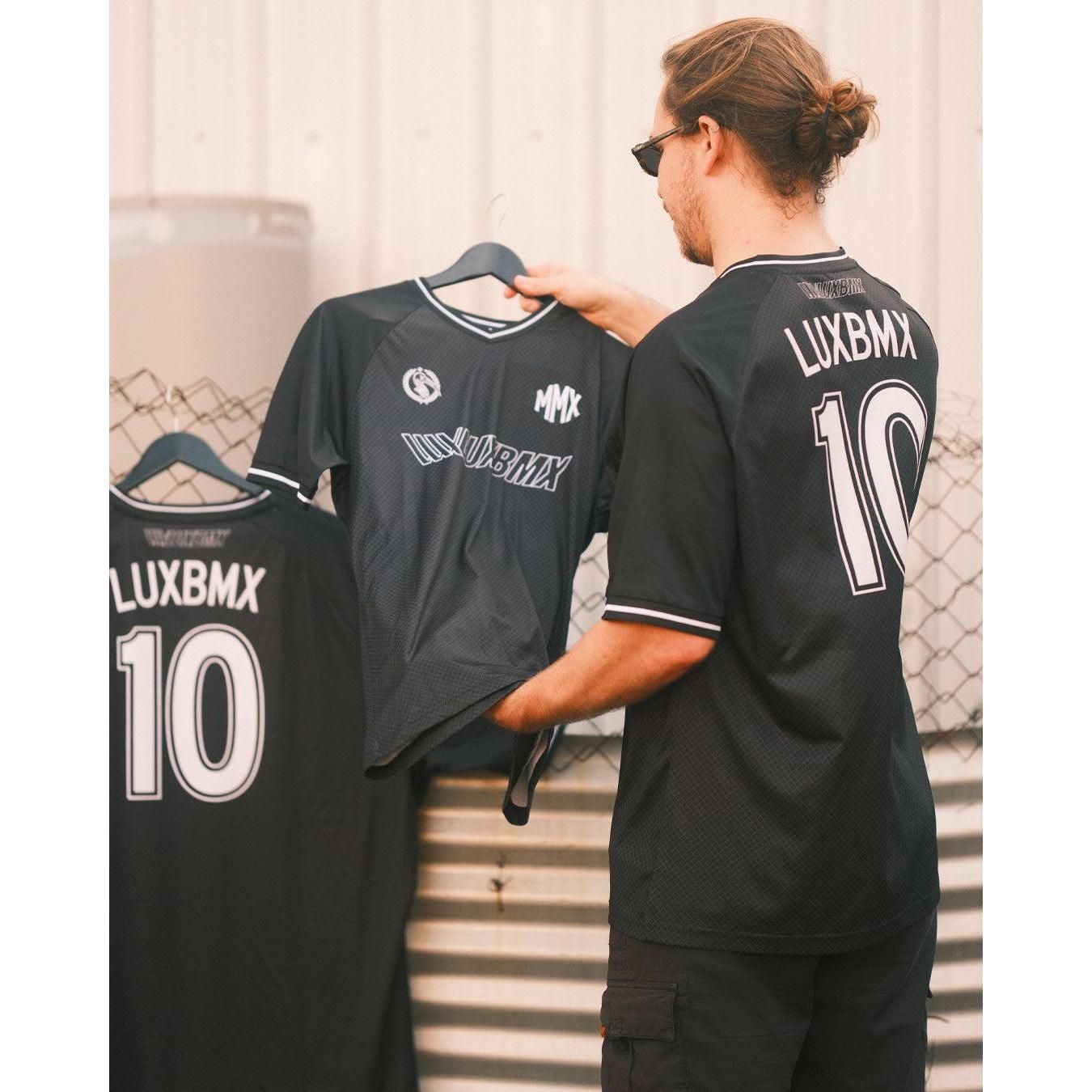 A man in sunglasses holds a black LUXBMX MMX Soccer Jersey with a custom back print (number 10), standing next to two similar breathable bike jerseys hanging on a fence.