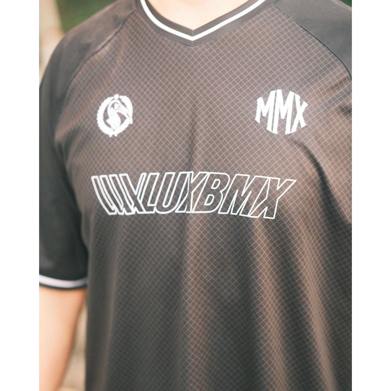 A person wears the LUXBMX MMX Soccer Jersey - Custom Back Print, featuring black, breathable fabric, "LUXLUXBMX" white text, "MMX" abbreviations, and a circular chest logo.