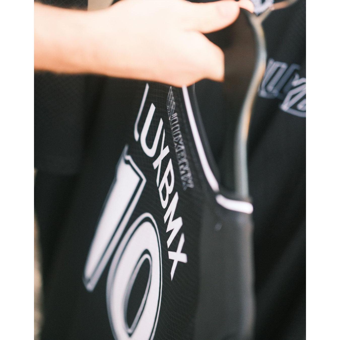 A person holds a black LUXBMX MMX Soccer Jersey - Custom Back Print on a hanger, highlighting its stylish look and breathable design perfect for BMX riders.
