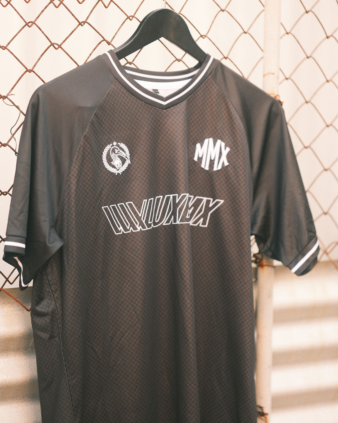 The LUXBMX MMX Soccer Jersey - Custom Back Print features a black and gray design with white trim and logos, displayed on a black hanger against a chain-link fence—perfect for those seeking a custom BMX jersey style.