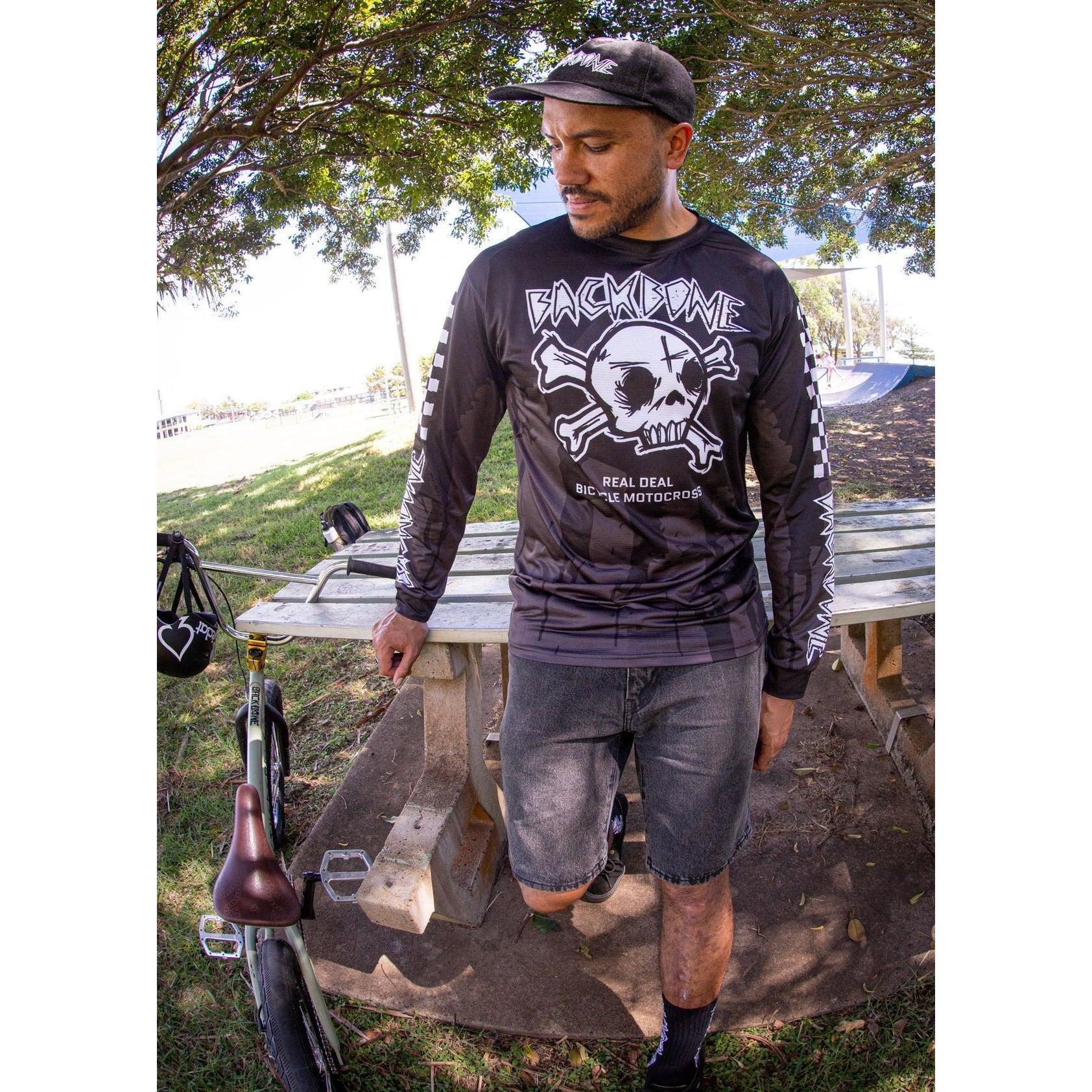 A man in a black Backbone 2026 Gnar Drip Jersey and cap stands outdoors near a picnic table with a BMX bike beside him.