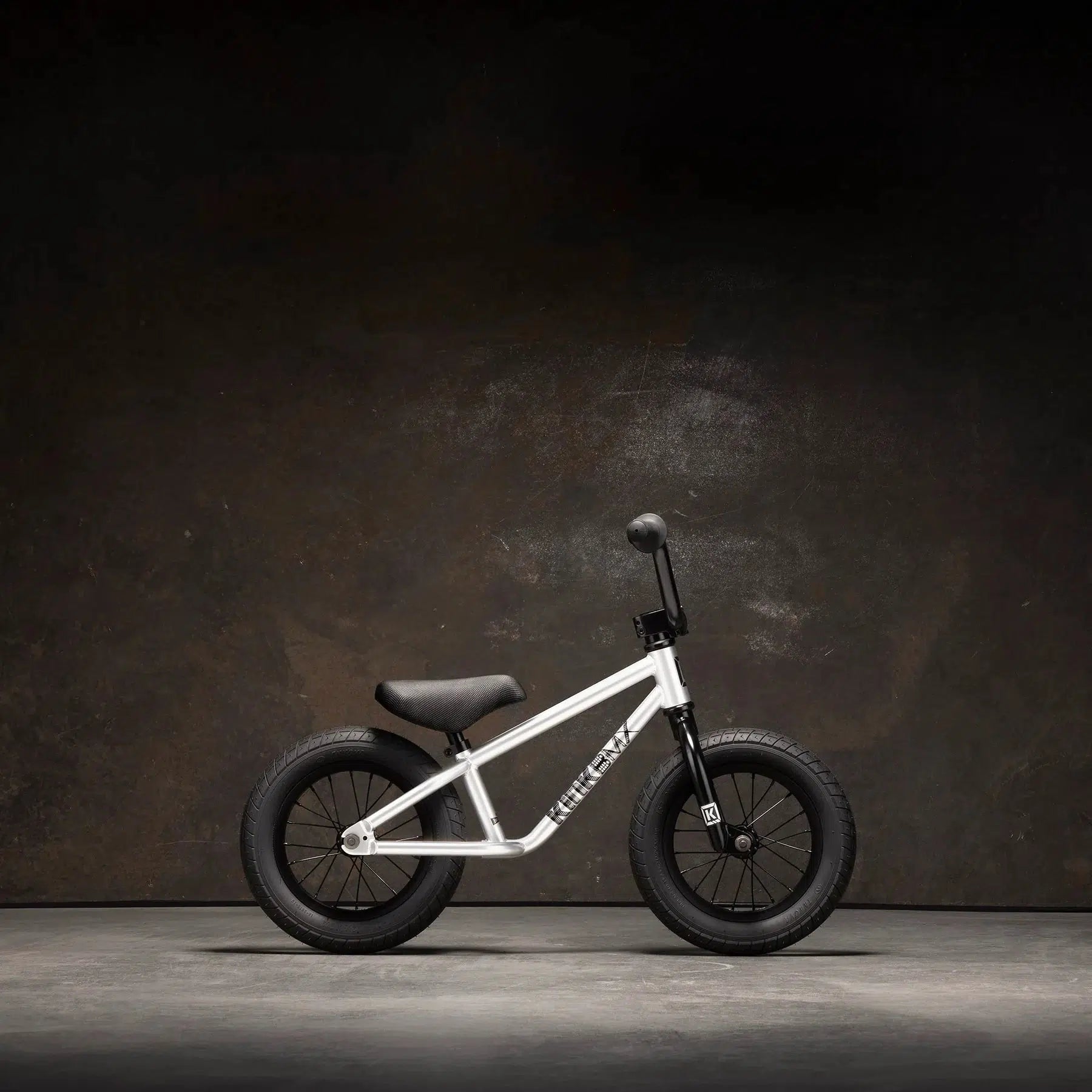 A small Kink Coast 12 Inch Balance Bike in silver, featuring black tires and seat, stands against a plain dark background.