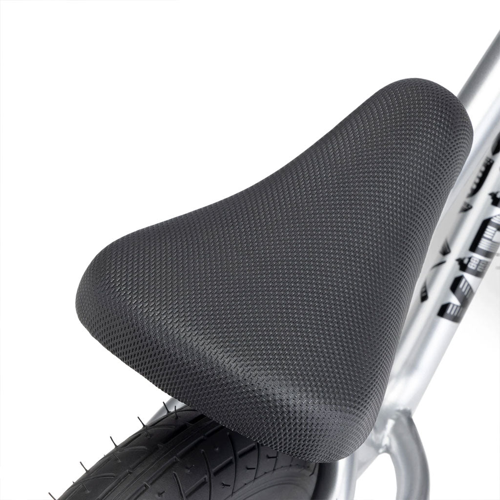 Close-up of a black textured bike seat on the silver frame of a Kink Coast 12 Inch Balance Bike, above a large black tire.