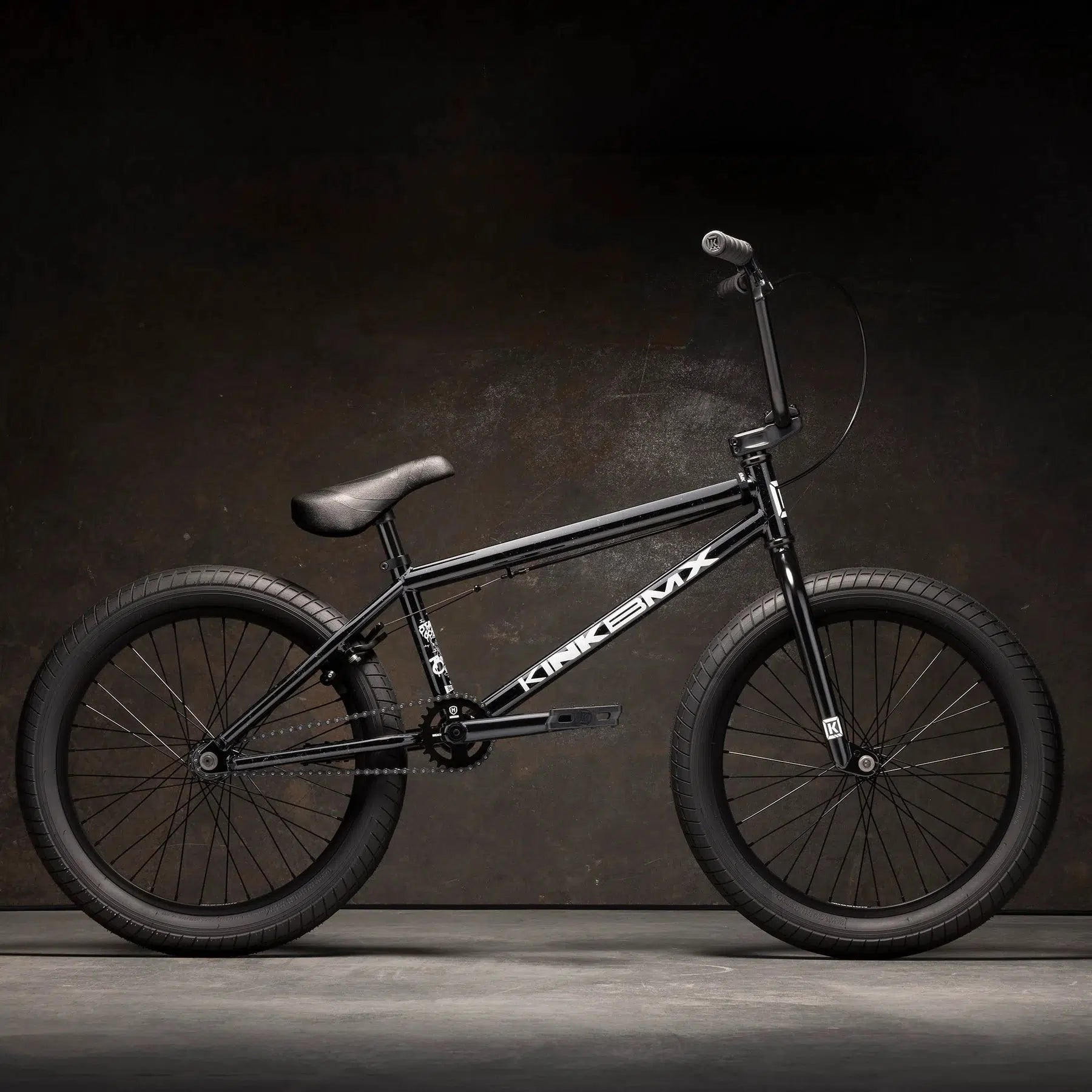 The Kink Curb 20 Inch Bike, in black, showcasing its sturdy frame and thick tires, is set against a dark textured background. It boasts a curved seat, Kink Ace Grips for enhanced control, and Mission Tracker Tires that ensure excellent traction on any terrain.