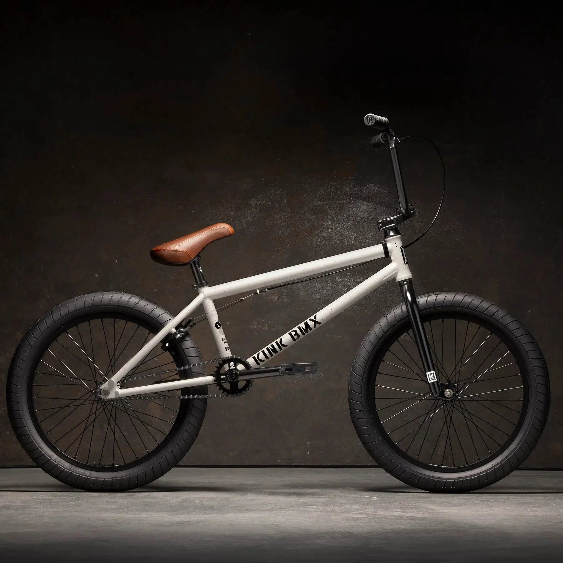 The Kink Gap 20 Inch Bike boasts a stylish white semi-chromoly frame, complemented by black wheels and a brown seat, making it an ideal choice for beginner riders against a dark backdrop.