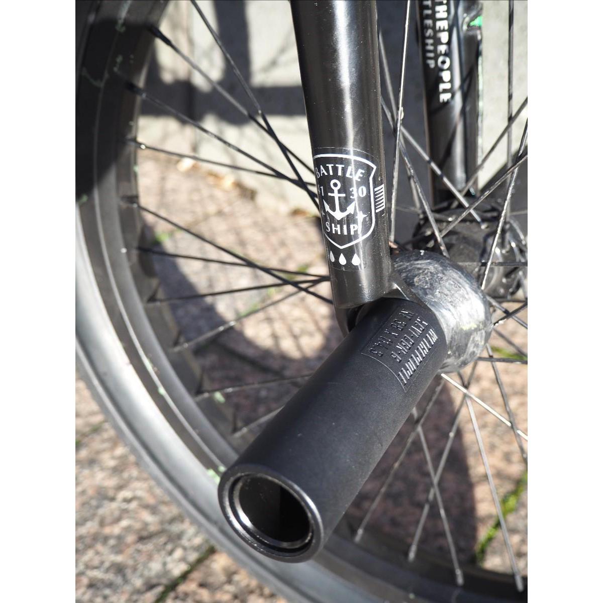 Close-up of a BMX bike front wheel with black spokes, a fork labeled "BATTLESHIP," and a pair of Wethepeople Temper Pegs, featuring a durable chromoly core, set against pavement and a concrete curb.