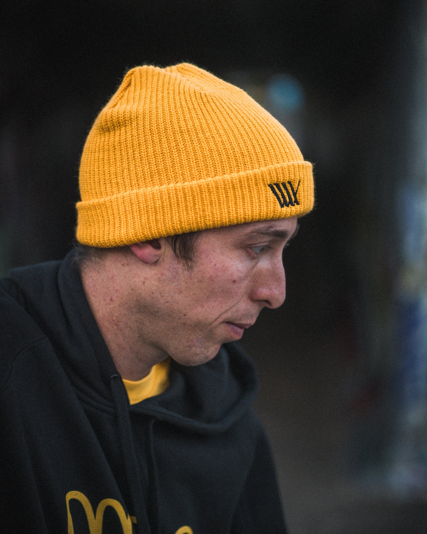 Wearing a yellow LUXBMX Flag Beanie and a black hoodie, a person is shown in profile against a dark, blurred background.