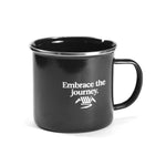The LUXBMX Journey Camp Mug is a black enamel-coated steel mug with a handle, featuring “Embrace the journey.” and a white mountain graphic printed on the side.