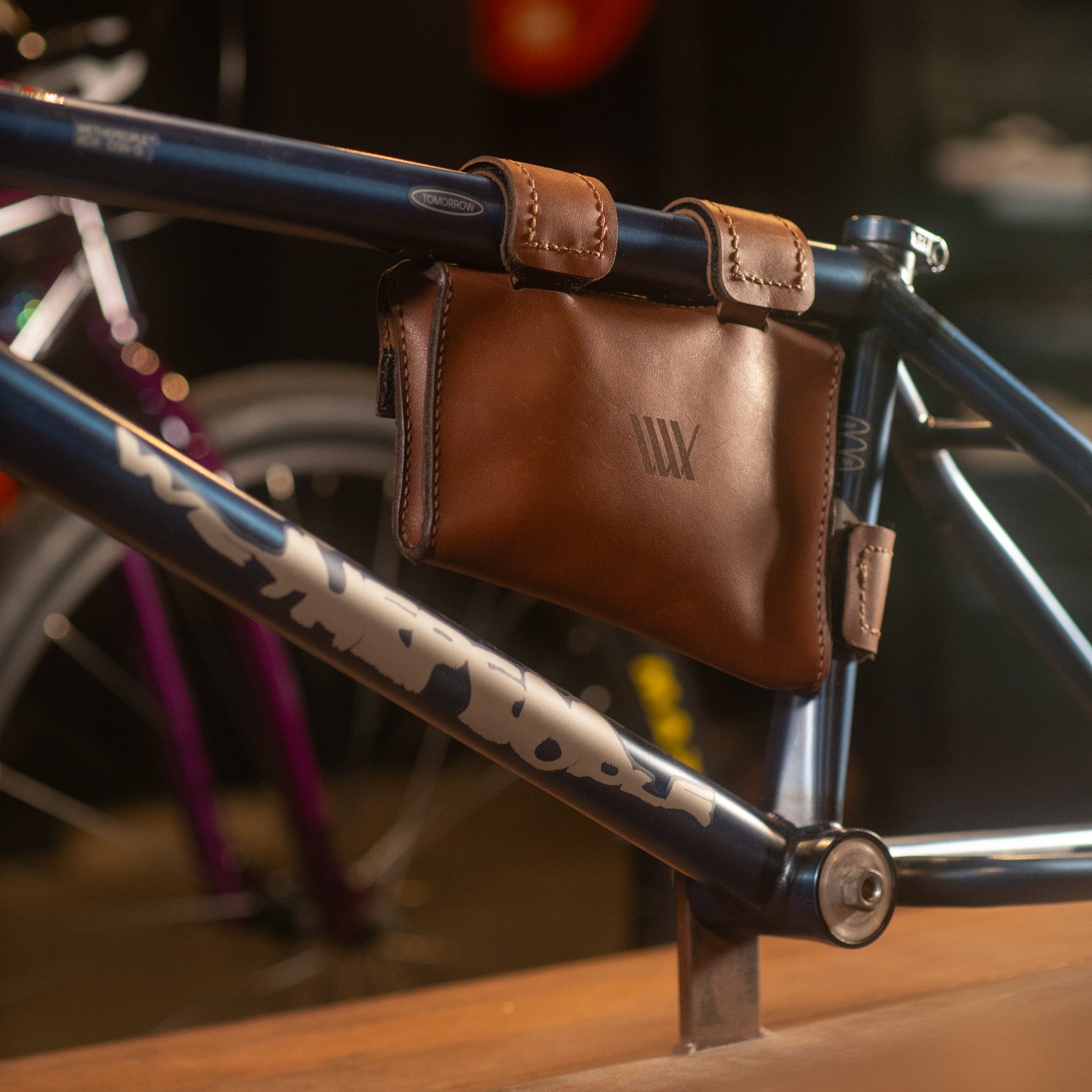 A LUXBMX Leather Frame Bag is fastened to a blue bike with leather straps. This handmade accessory adds style, and part of another bicycle appears in the background.