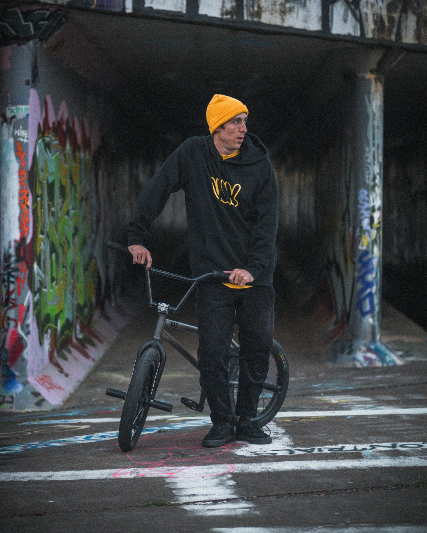 A person in a yellow beanie and the LUXBMX Throwie Hoodie - Black stands beside a BMX bike on a graffiti-covered path under a bridge.