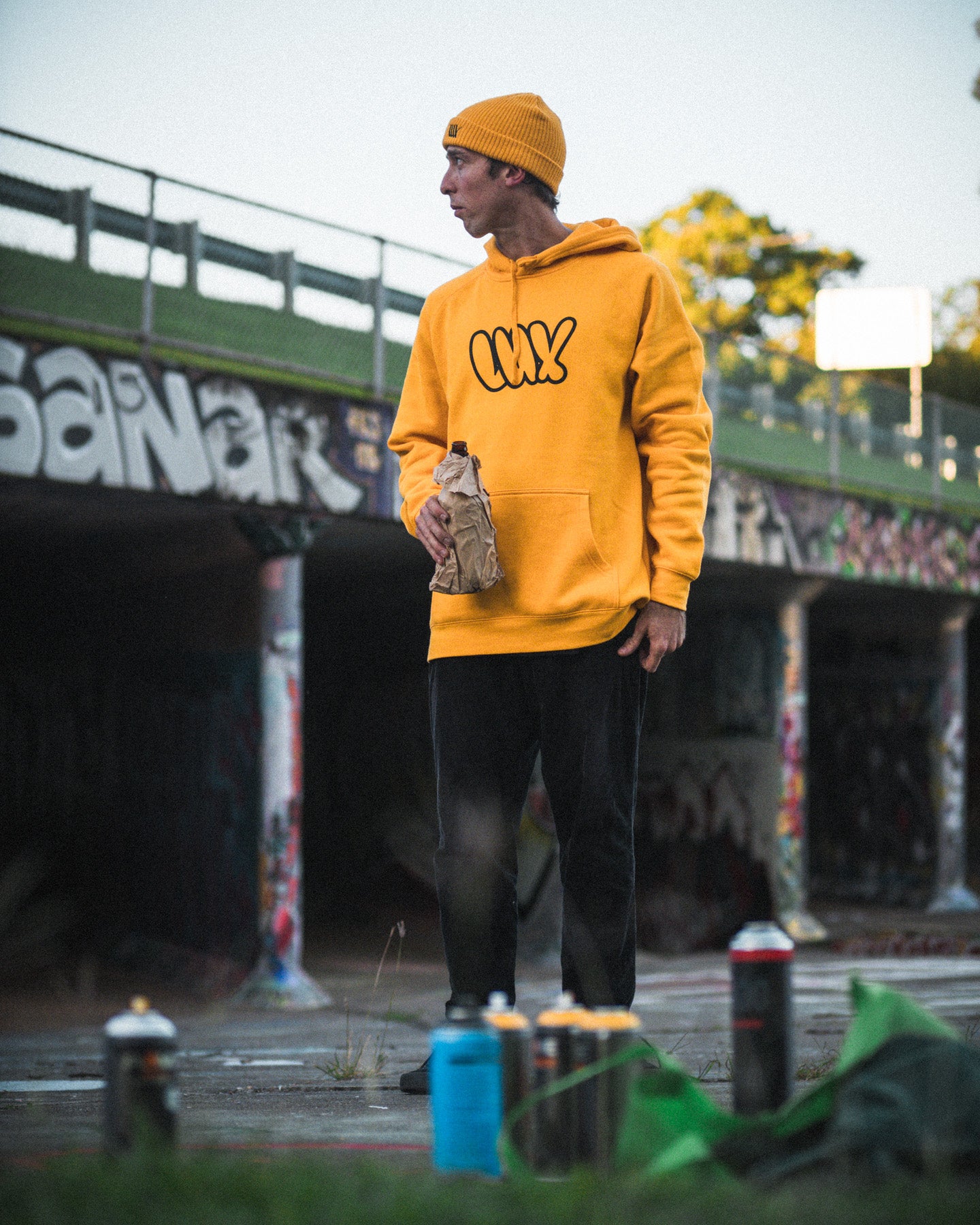 Wearing a yellow LUXBMX Throwie Hoodie, a person stands outdoors by a graffiti wall, holding a paper bag with spray paint cans at their feet.