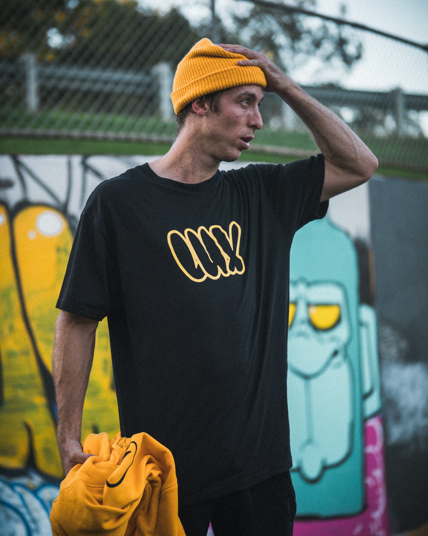 A man in a yellow beanie and the black LUXBMX Throwie Tee stands before a colorful graffiti wall, holding a yellow sweatshirt. The midweight cotton tee features a bold screenprinted graphic.