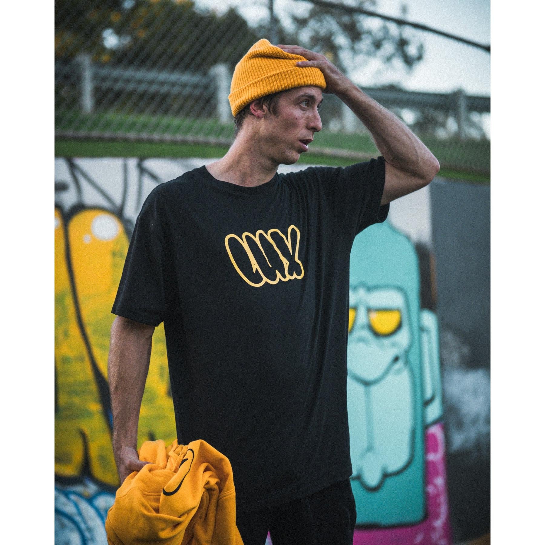A man in a yellow beanie and the black LUXBMX Throwie Tee stands before a colorful graffiti wall, holding a yellow sweatshirt. The midweight cotton tee features a bold screenprinted graphic.
