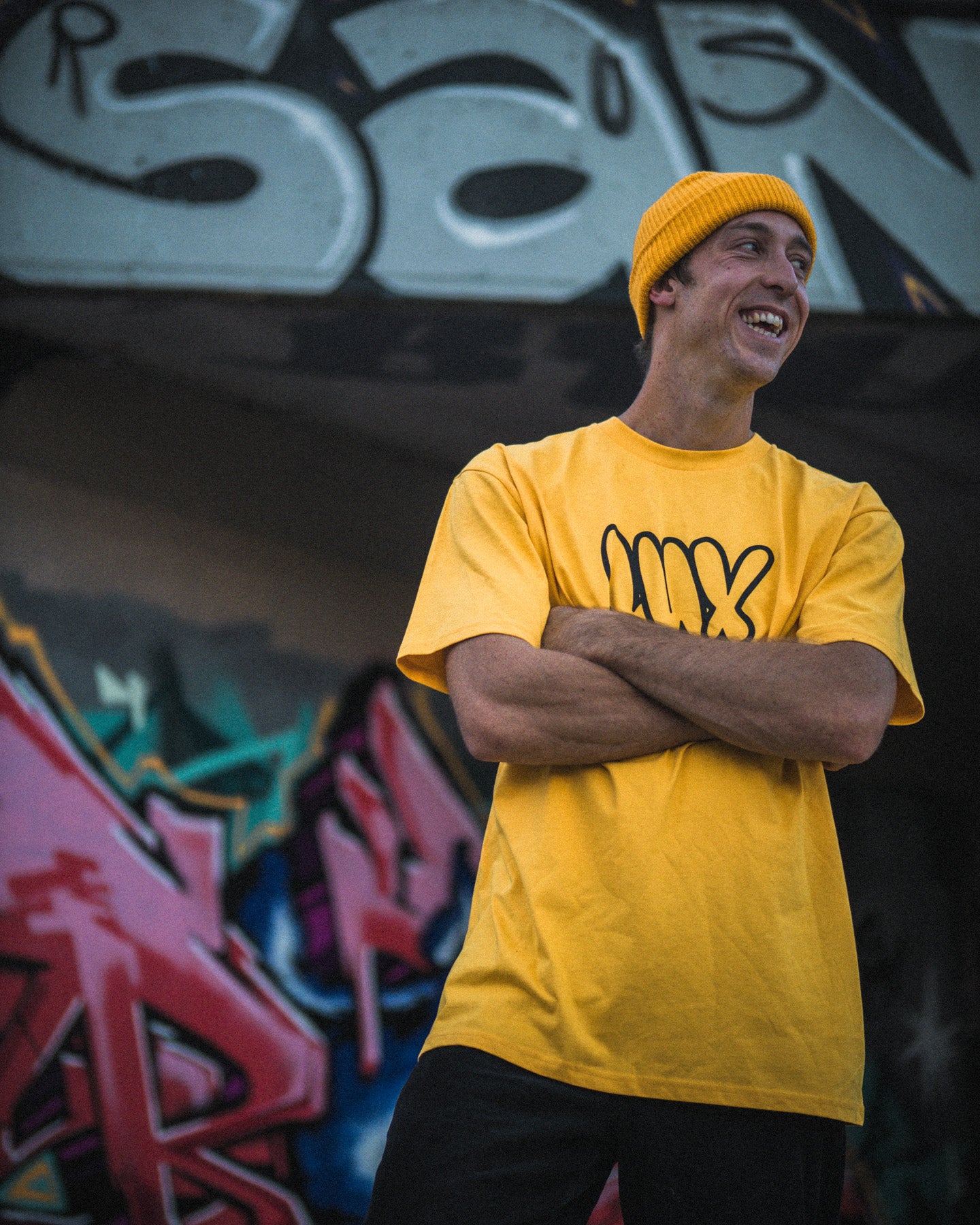 Smiling with arms crossed, a person wears the yellow LUXBMX Throwie Tee, featuring a throwie-style logo, while standing in front of a colorful graffiti wall.
