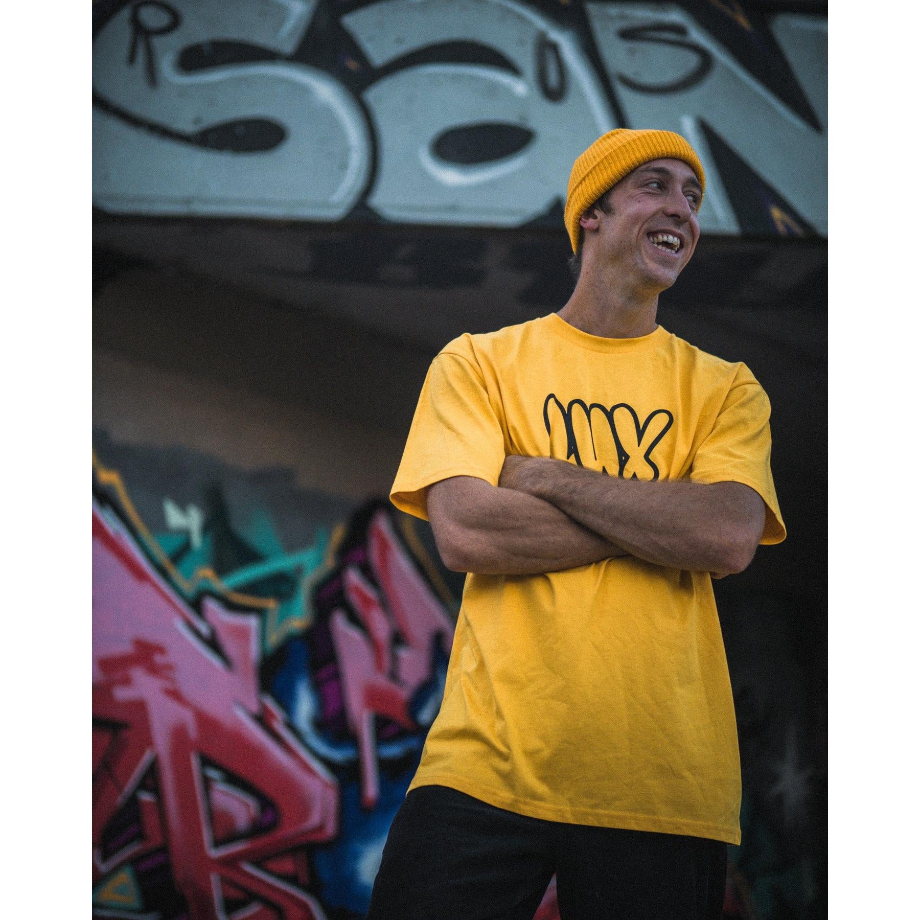 Smiling with arms crossed, a person wears the yellow LUXBMX Throwie Tee, featuring a throwie-style logo, while standing in front of a colorful graffiti wall.