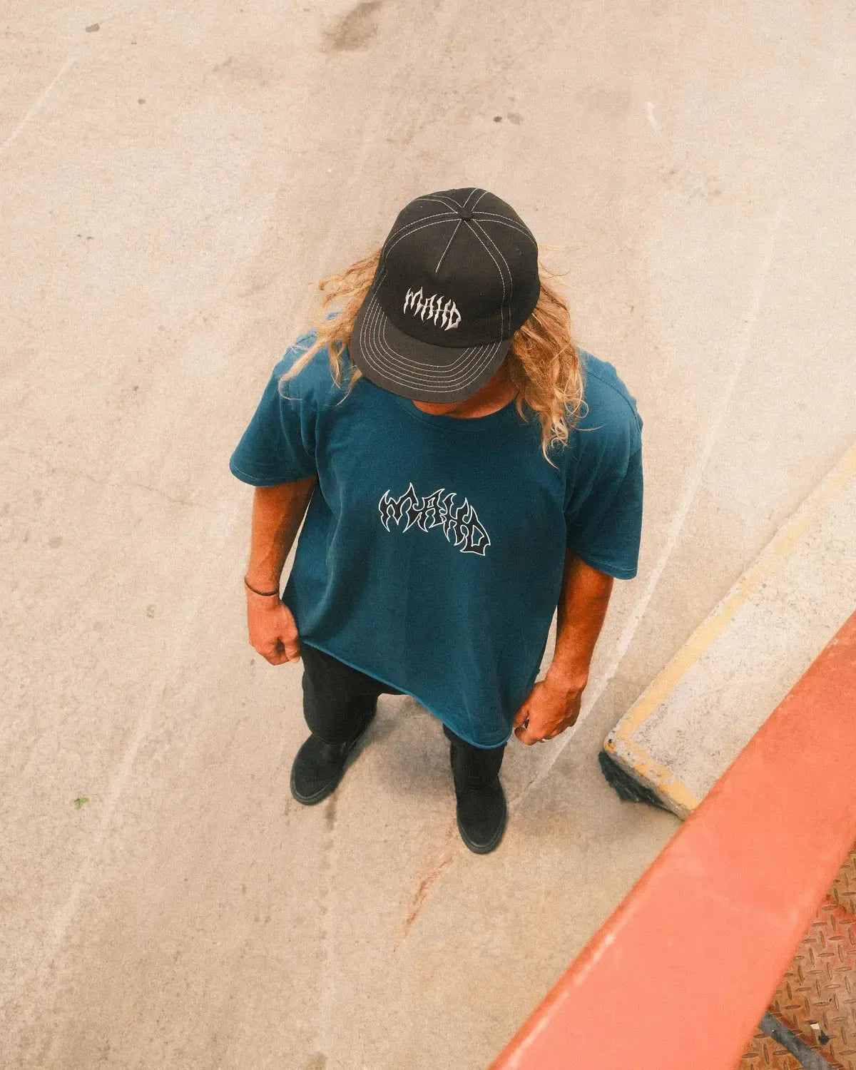 A person with long hair wearing a black cap, sand colour Mahd Metal Tee with centre logo, black pants, and black shoes stands on a concrete surface, viewed from above.