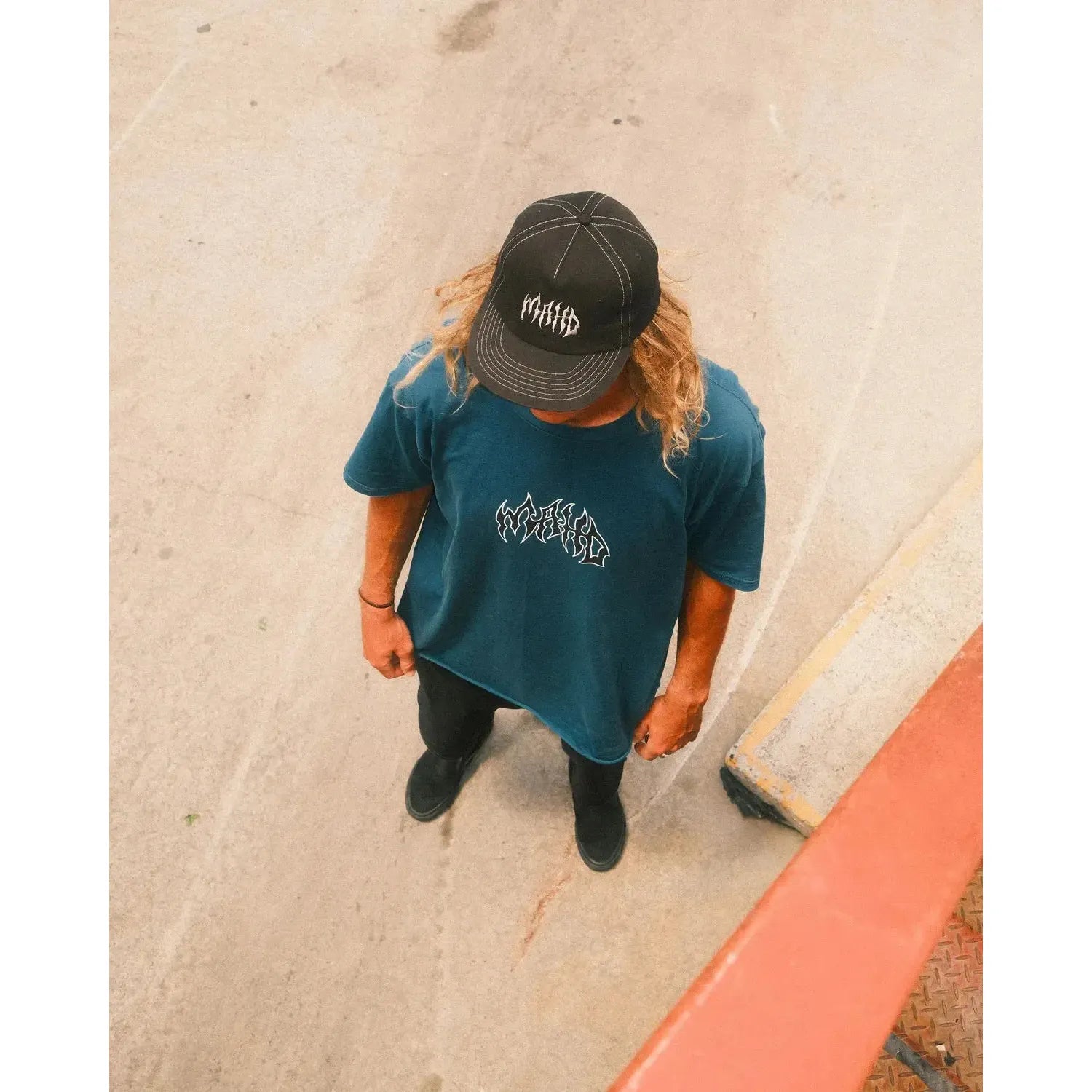 A person with long hair wearing a black cap, sand colour Mahd Metal Tee with centre logo, black pants, and black shoes stands on a concrete surface, viewed from above.