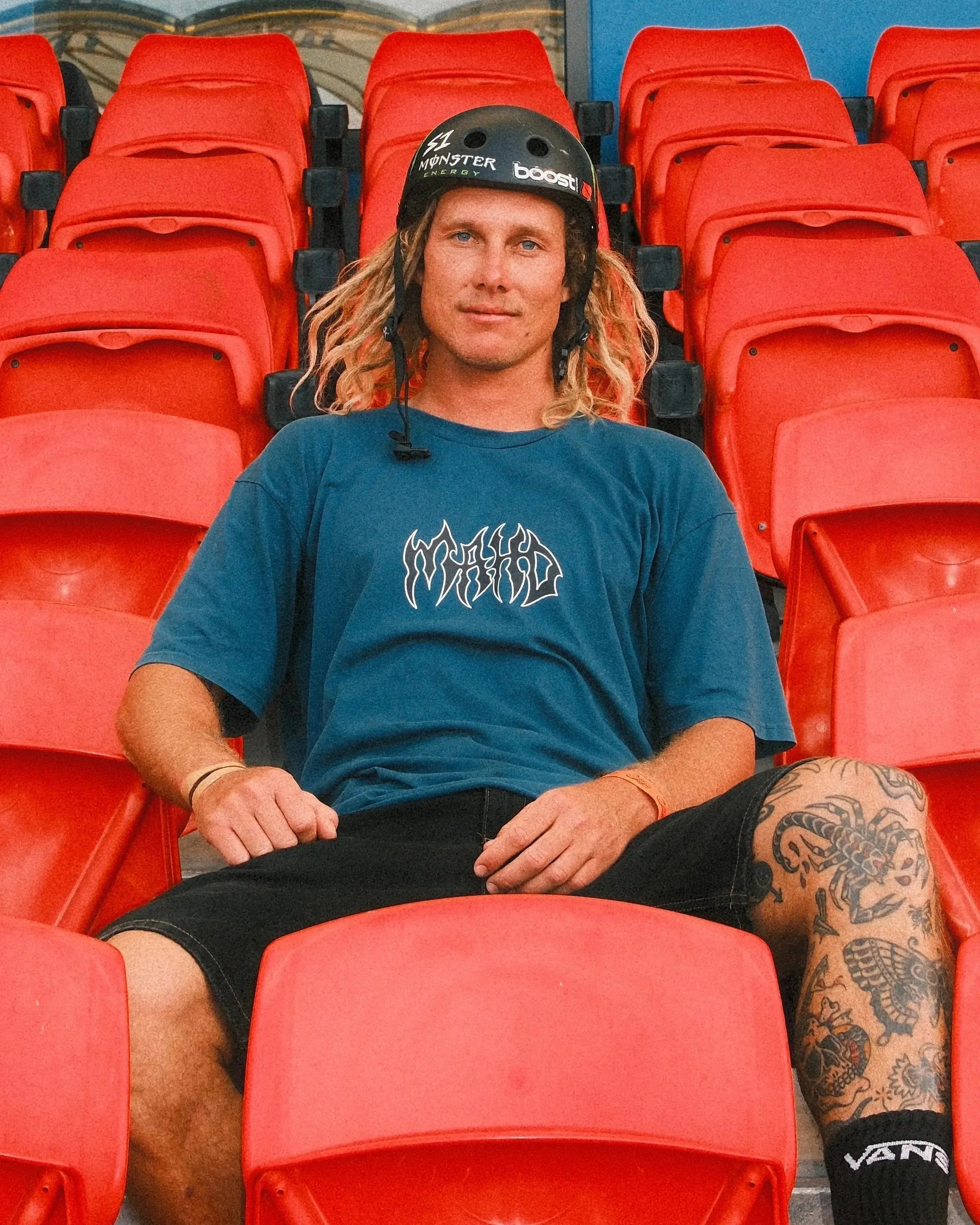 A person with a helmet and dreadlocks sits on red stadium seats, wearing the blue Mahd Metal Tee with a centre logo, black shorts, and showcasing a leg tattoo.