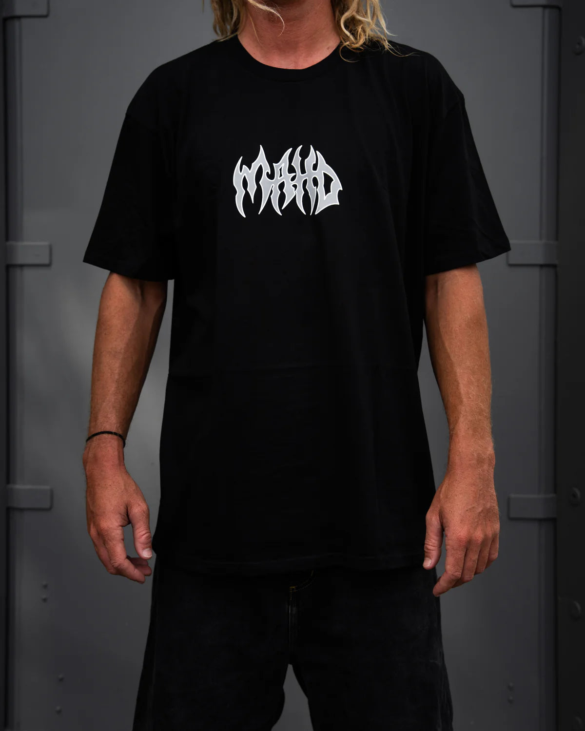 A person wearing the Mahd Metal Tee in sand with a white center logo stands against a dark gray background.