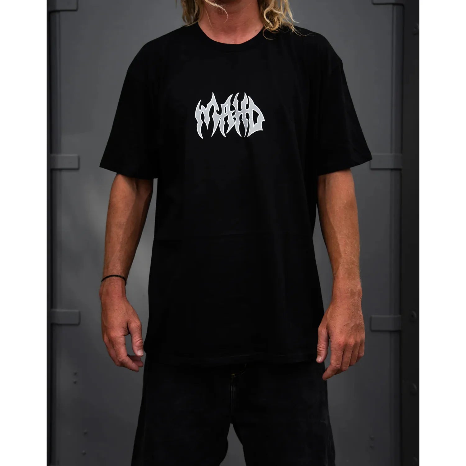 A person wearing the Mahd Metal Tee in sand with a white center logo stands against a dark gray background.