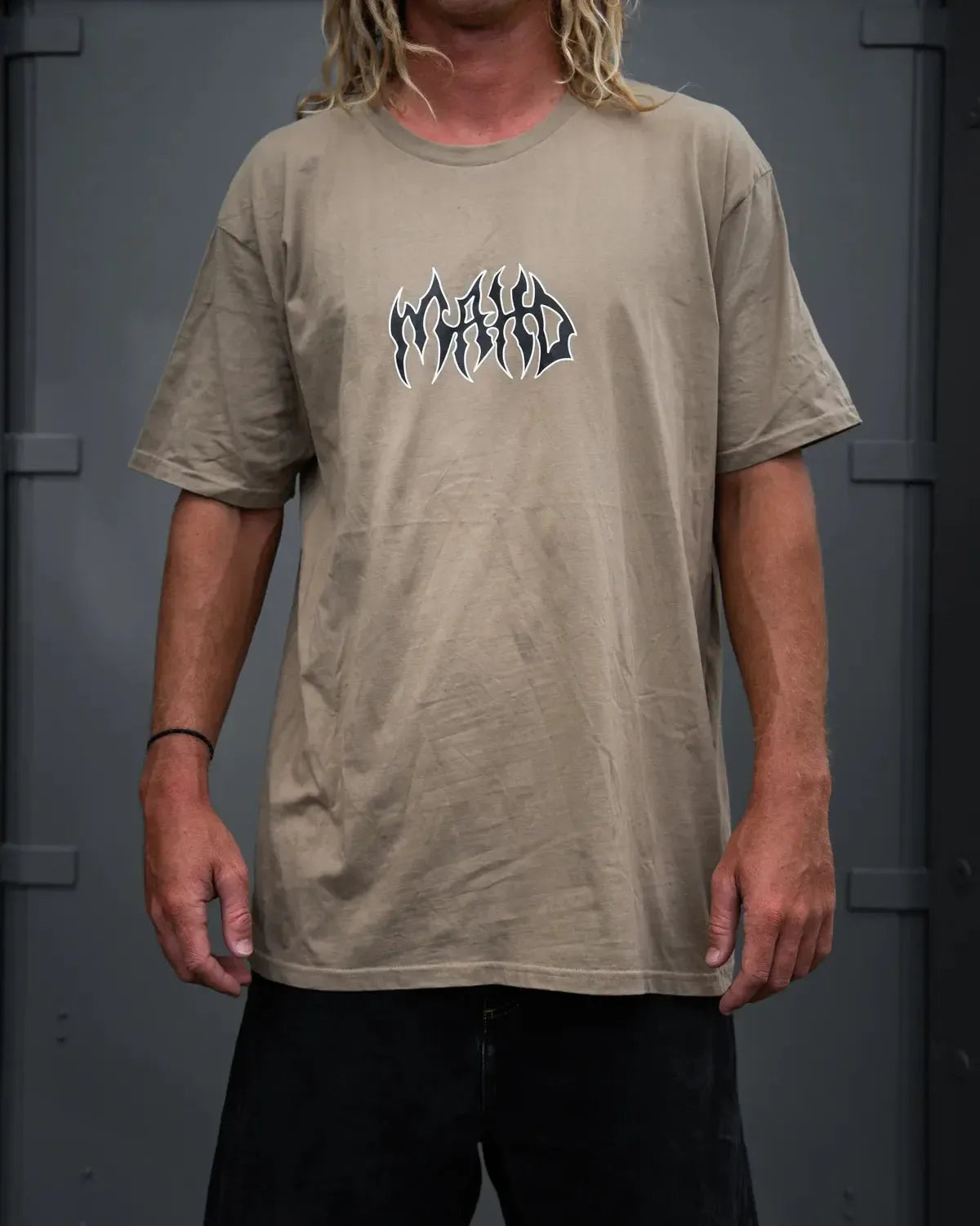 A person wears a sand Mahd Metal Tee featuring a central "World World" logo, standing in front of a gray metal background.