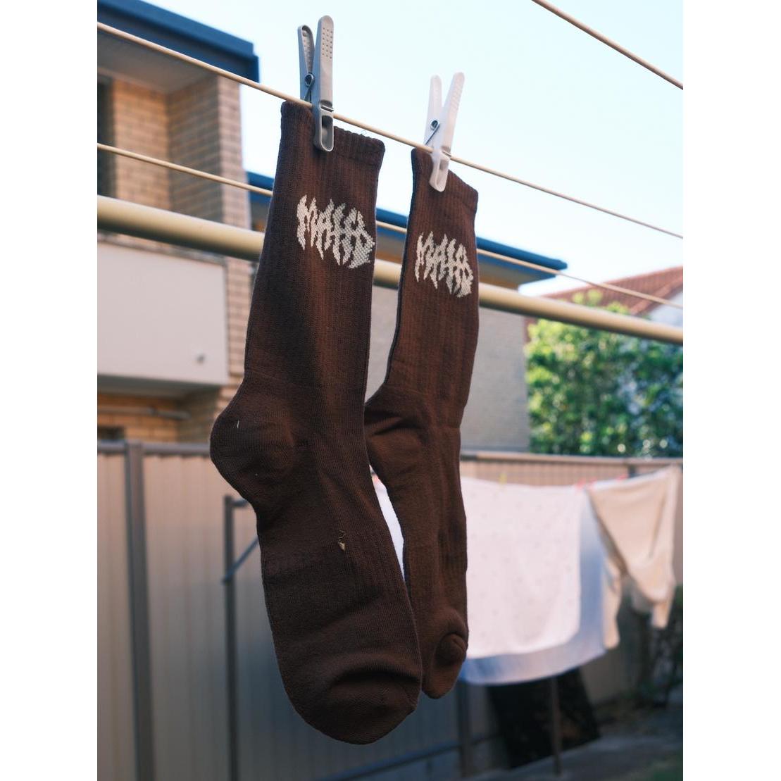 A pair of Mahd Metal Socks, brown with white designs, hang from a clothesline by clothespins. Made from a bamboo blend, these one-size-fits-most socks offer everyday comfort against a backdrop of laundry in a suburban backyard.