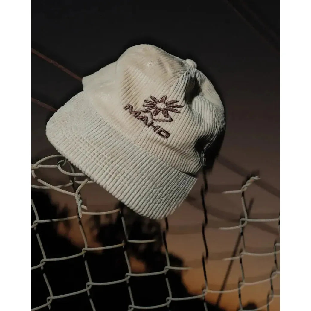The Mahd Botanical Corduroy Hat, featuring cords and an embroidered "MANTO" logo, sits on a wire fence at dusk, delivering Boyd Hilder-inspired style.