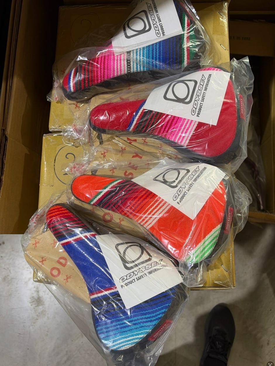 Four Odyssey Mexican Blanket Railed Seats, featuring a multicolored design, are stacked in clear plastic packaging on top of cardboard boxes on a concrete floor.