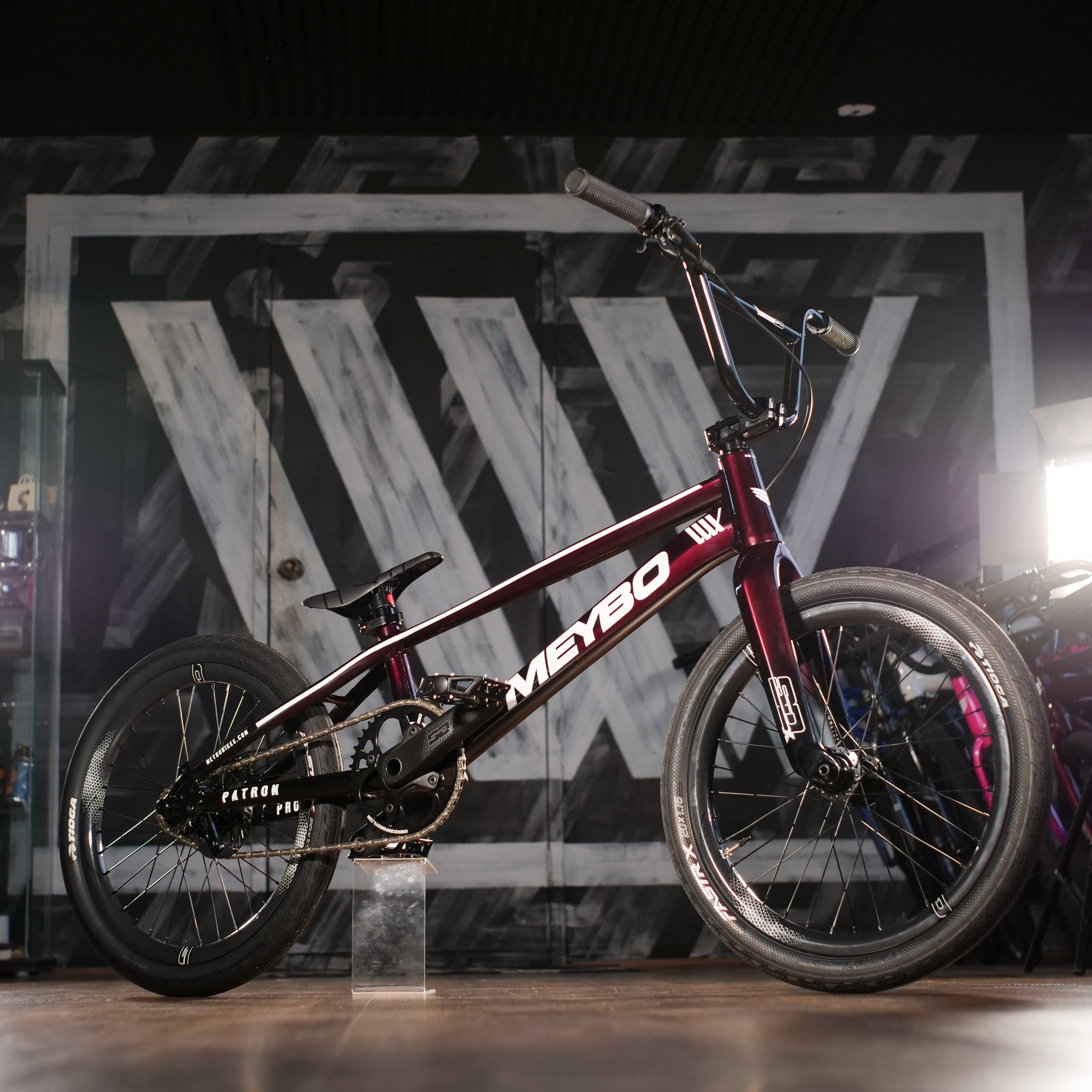 A Meybo 2025 Patron Pro XXL Bike in maroon with "MEYBO" branding and SD carbon rims is displayed indoors against a geometric black-and-white wall.