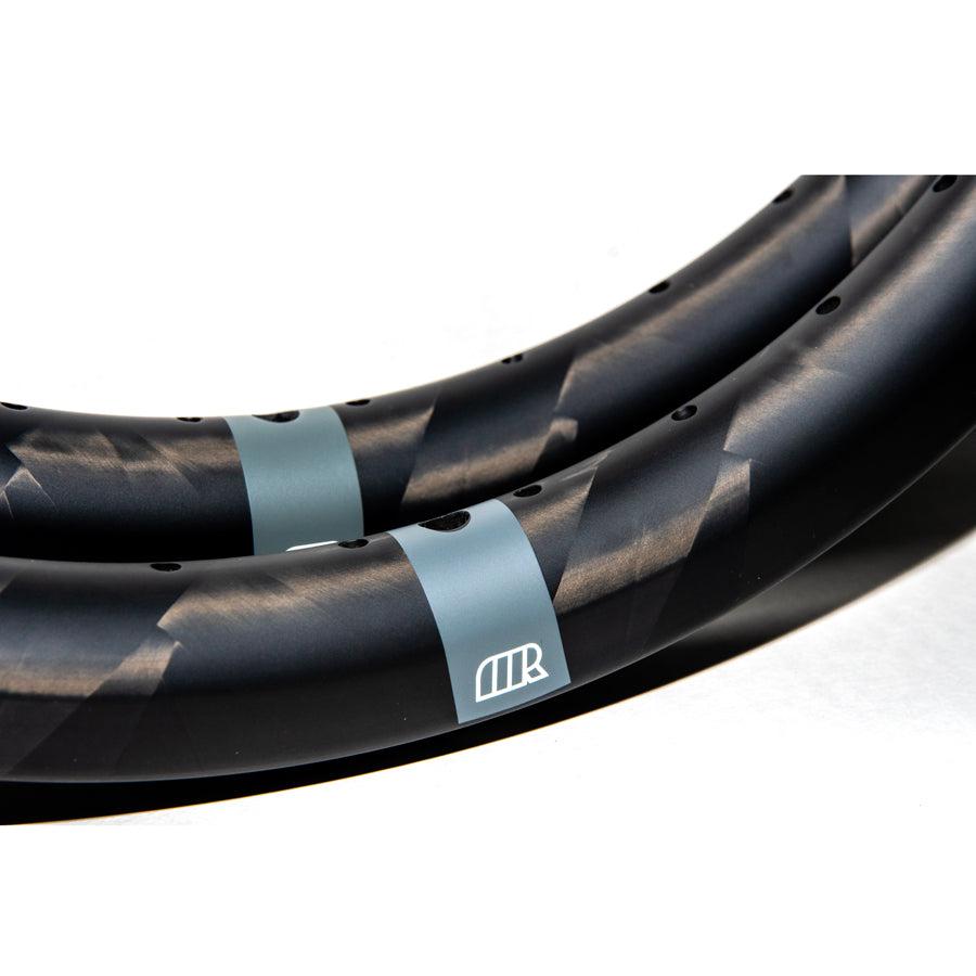 Close-up of a Michram OWT Carbon Rim Plain Logo (28 Hole), featuring a black matte finish with a geometric pattern, crafted from hand-woven T800 carbon fiber, and displaying a logo in a contrasting lighter color.