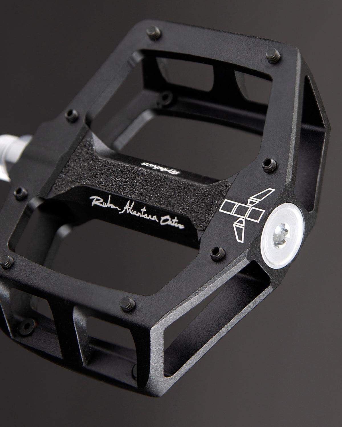 Close-up of a sleek black Fly Bikes Ruben Aluminum pedal with a metallic body and CNC Chromoly axle, featuring precision bearings. The visible branding stands out against the dark background.