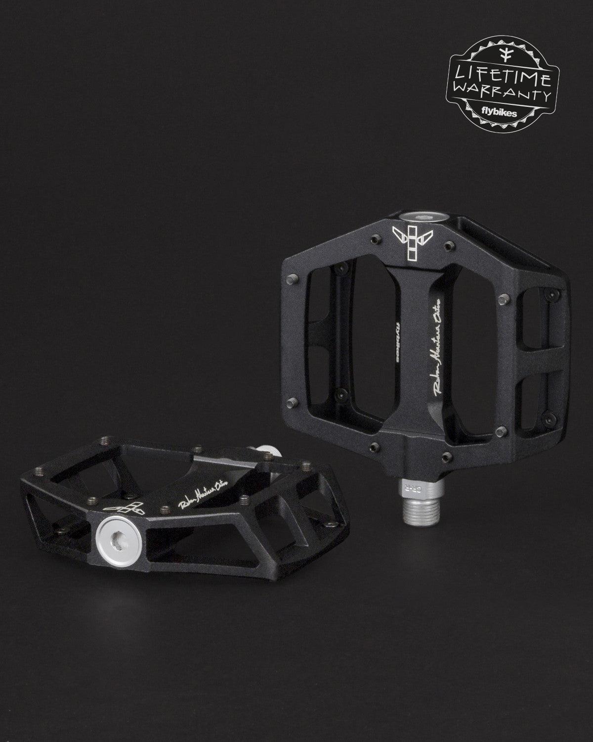 Two black Fly Bikes Ruben Aluminum Pedals with silver detailing are set against a dark backdrop. One is upright, highlighting its CNC Chromoly axle. The other is flat. A "Lifetime Warranty" logo in the top right promises durability with precision bearings.