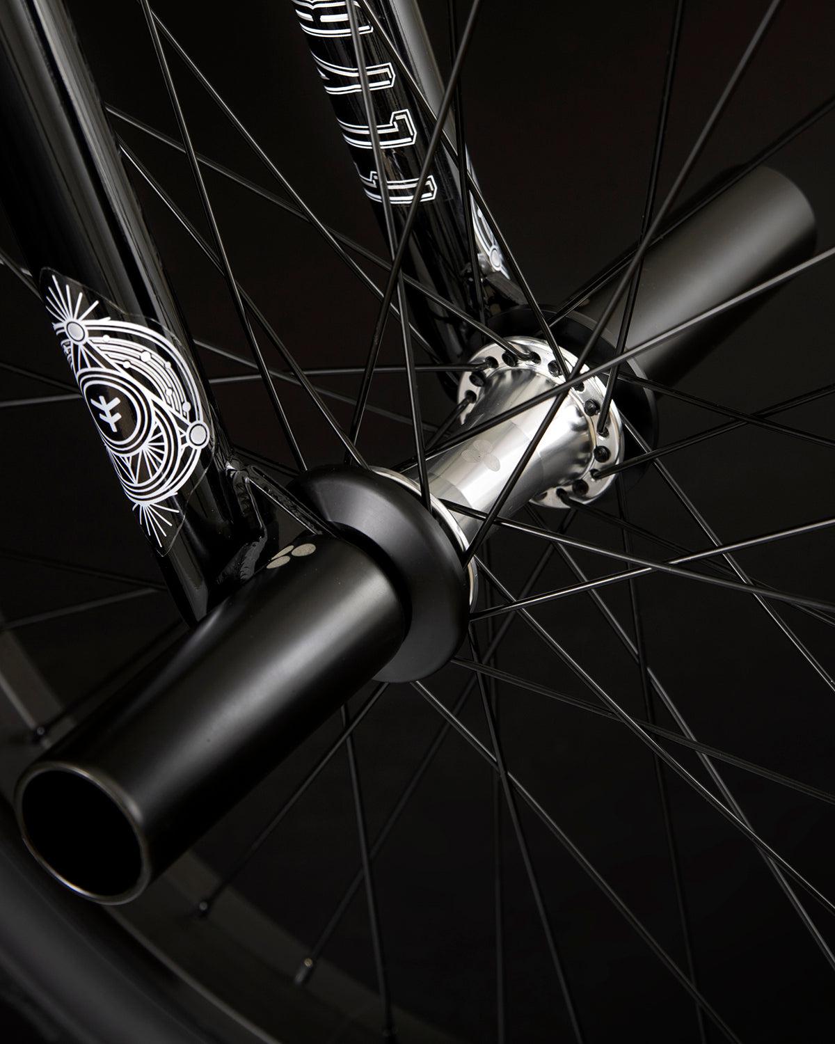 Close-up view of the Fly Bikes Proton FC 20 Inch Bike's black wheel, featuring an intricate spoke design, sealed hubs, and axle pegs, set against a dark background.