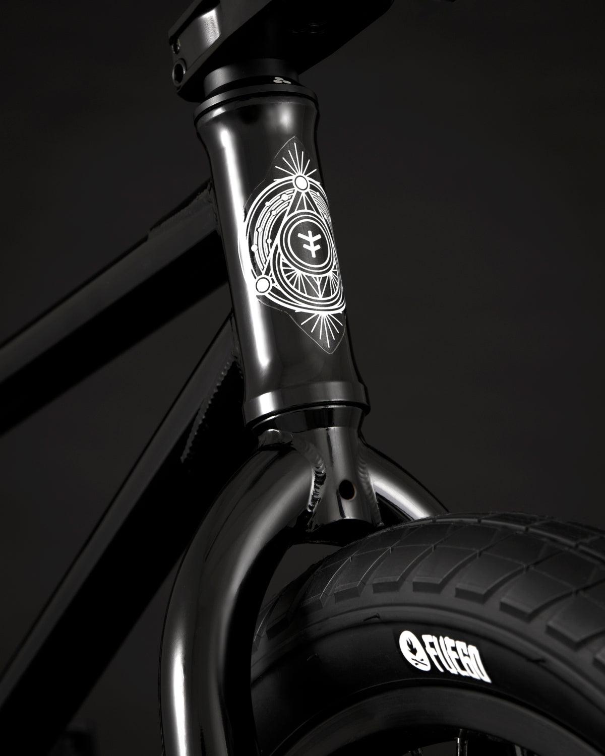 Close-up of the Fly Bikes Proton FC 20 Inch Bike, showcasing its black BMX design with a decorative emblem on the front. The bike features branded "Fiction" tires for added style. Its CrMo frame enhances durability, and the sealed hubs guarantee smooth rides against the dark backdrop.