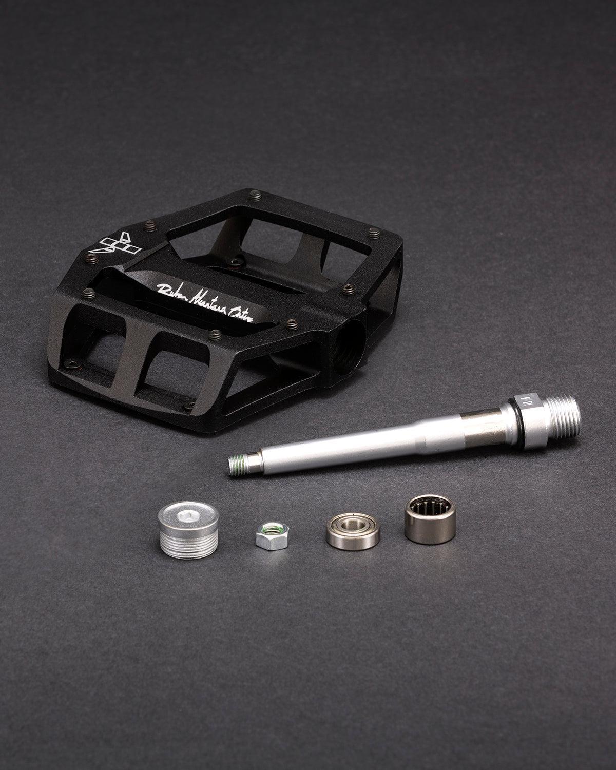 The black Fly Bikes Ruben Aluminum pedal is disassembled, revealing its components: an aluminum pedal body, CNC Chromoly axle, precision bearings, and hardware set against a dark background.