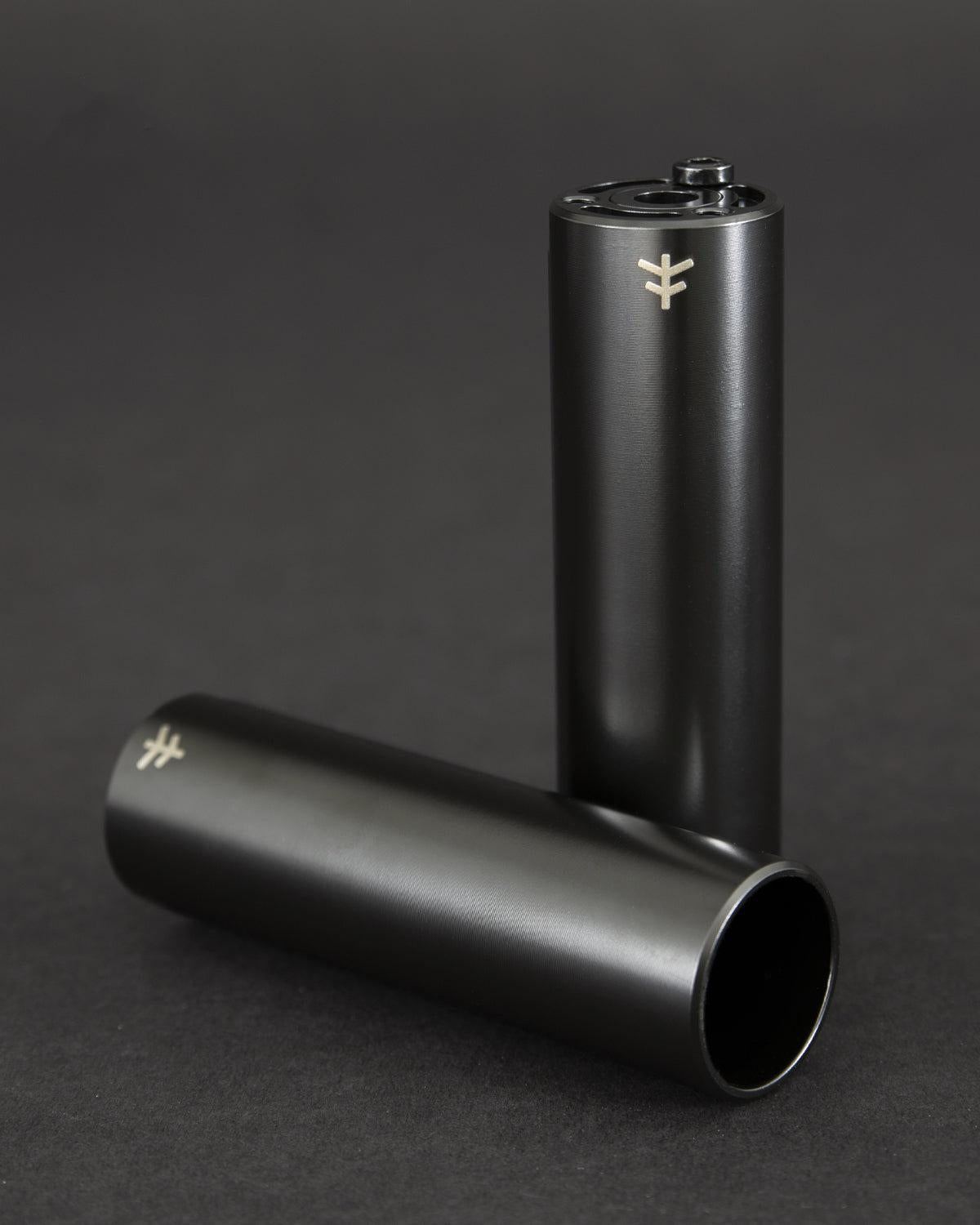 Two Fly Bikes Acero ST Pegs in black rest on a dark surface; one stands upright, showcasing its sleek design reminiscent of CNC machined 4140 Cr-Mo precision, while the other lies down with an air of sophistication.